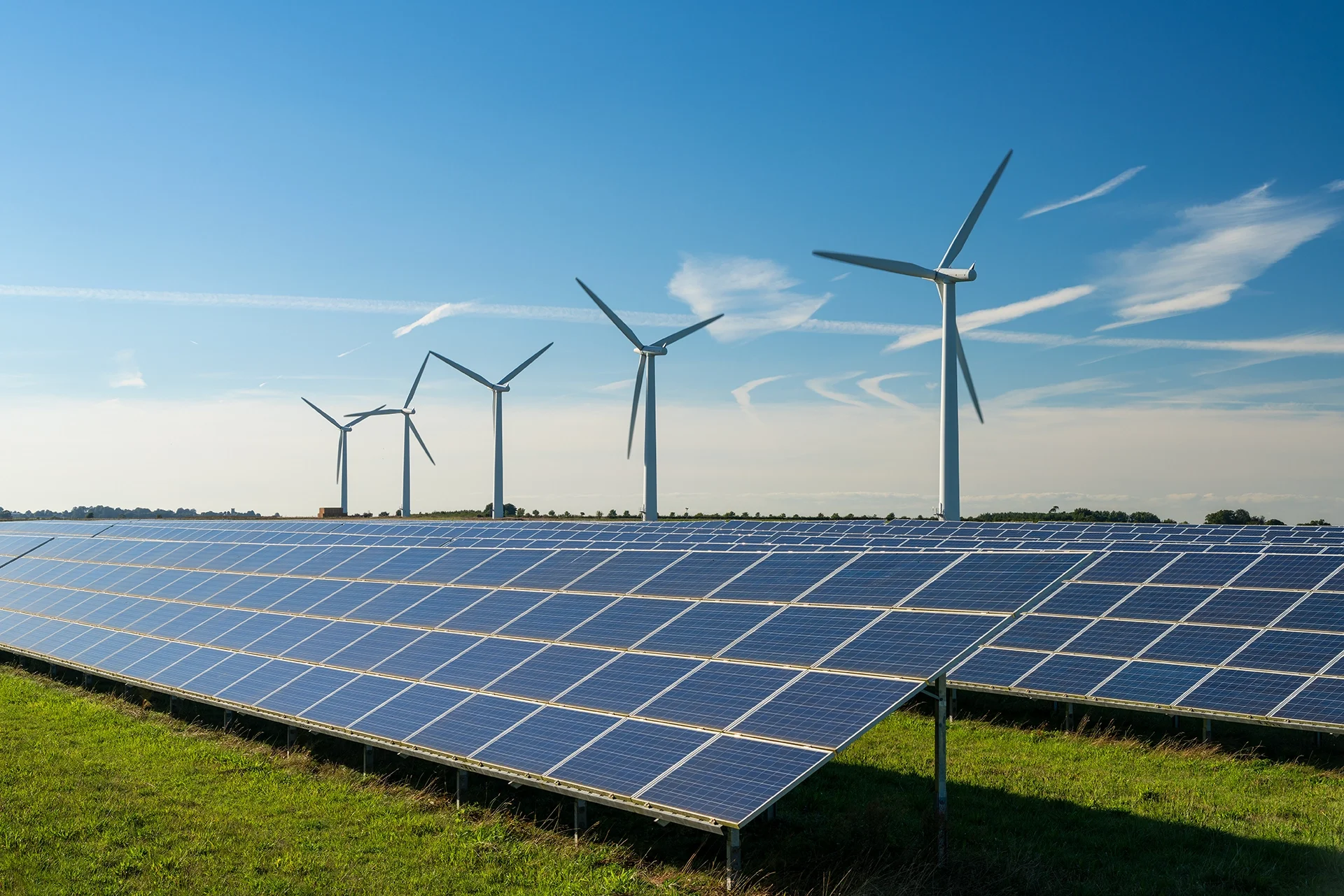 Solar panels in the foreground with wind turbines in the background under a blue sky with wispy clouds.