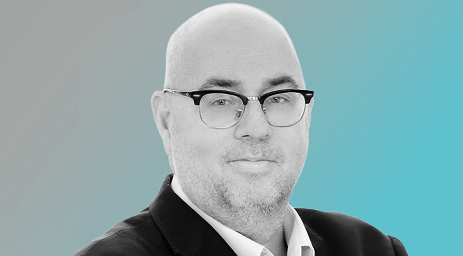 Black and white portrait of a man with glasses, balding head, wearing a suit jacket and white shirt, against a gradient blue background.