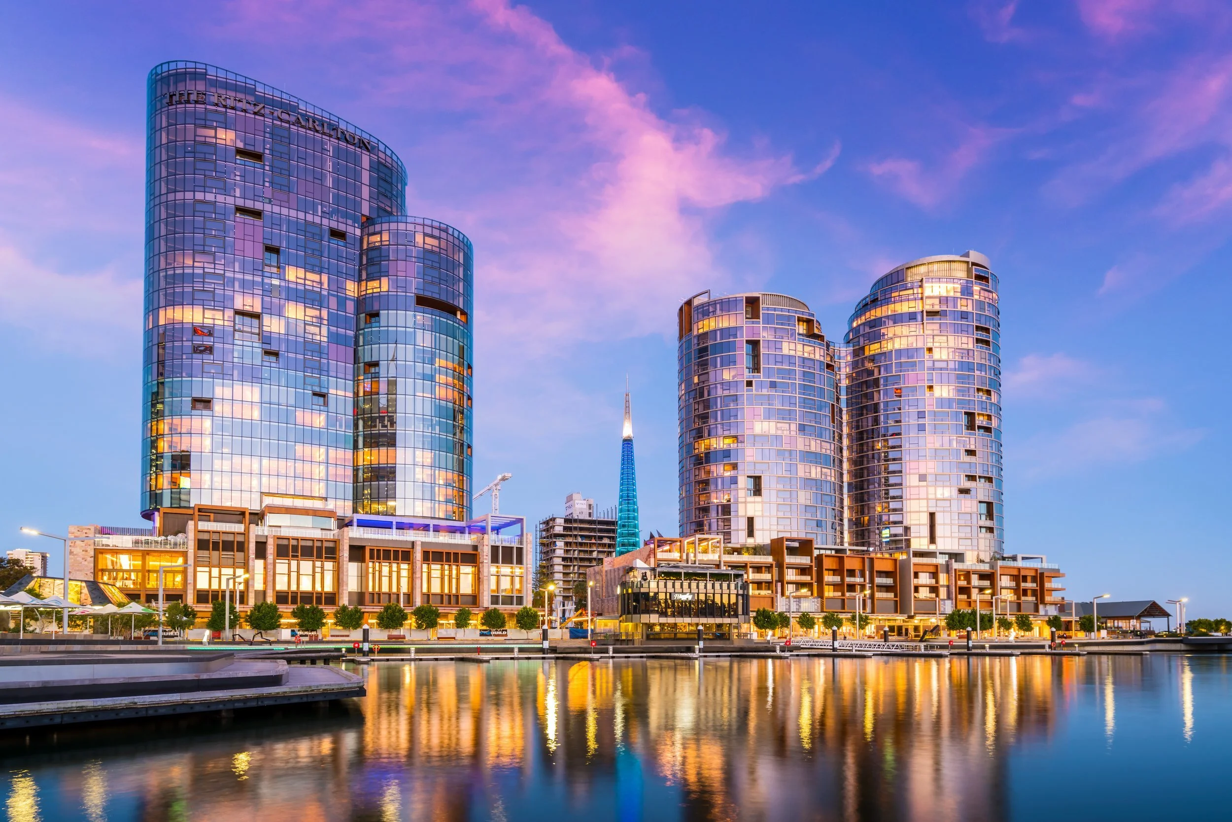 City skyline at sunset with modern glass buildings reflecting the sky and water in the foreground.