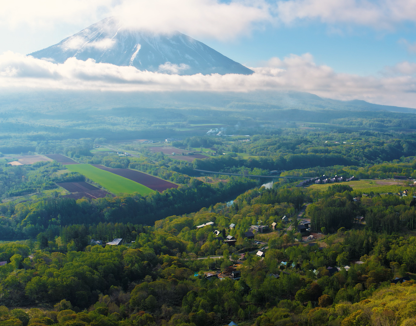 Scenic view of a mountain partially covered by clouds, with lush green fields and a small village in the foreground.