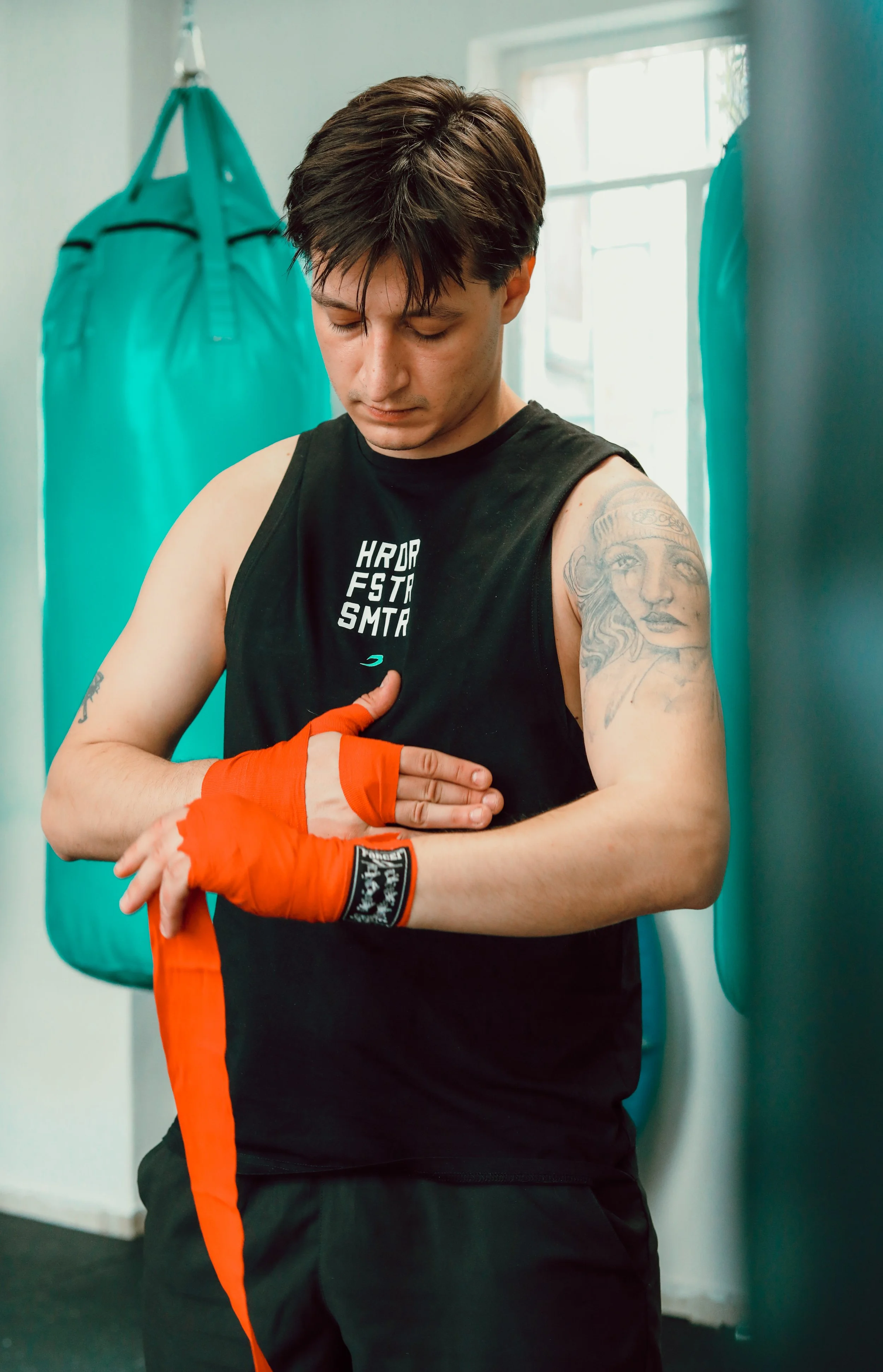 Two men training in a gym, one wearing boxing gloves and the other holding focus mitts, against a teal wall.