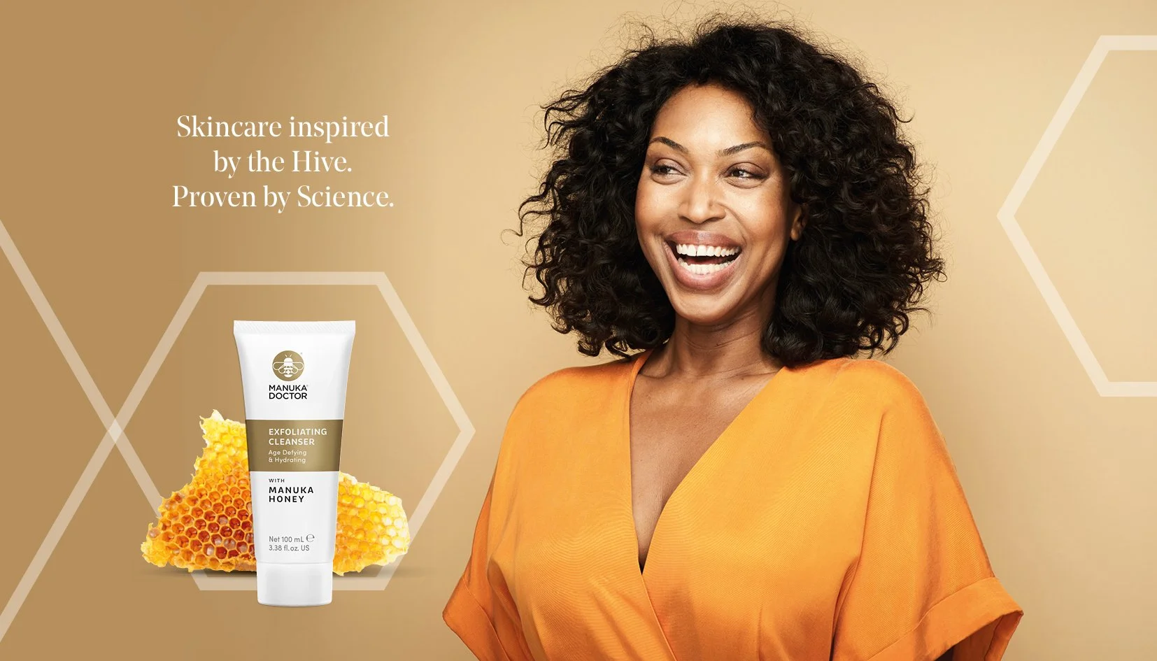 A woman with curly black hair smiling, wearing an orange top. To her left is a tube of Manuka Doctor exfoliating cleanser with honey and honeycomb images behind it. The background is beige with hexagonal patterns. Text reads: 'Skincare inspired by the Hive. Proven by Science.'