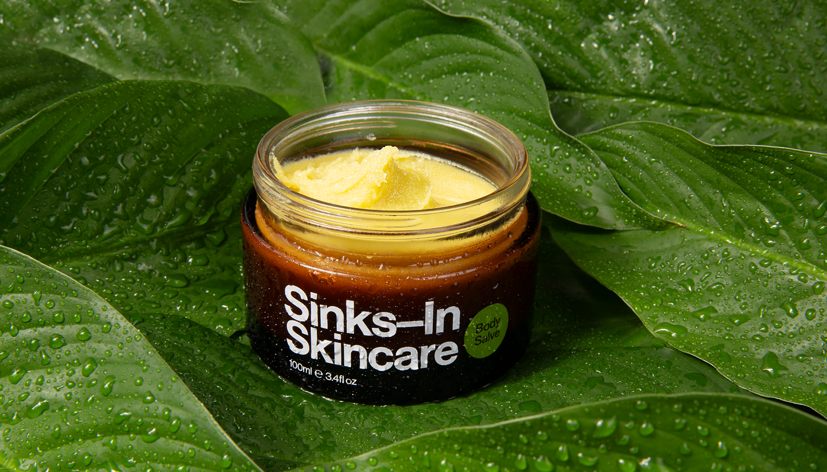 Open jar of body salve labeled 'Sinks In Skincare' resting on large green leaves with water droplets.