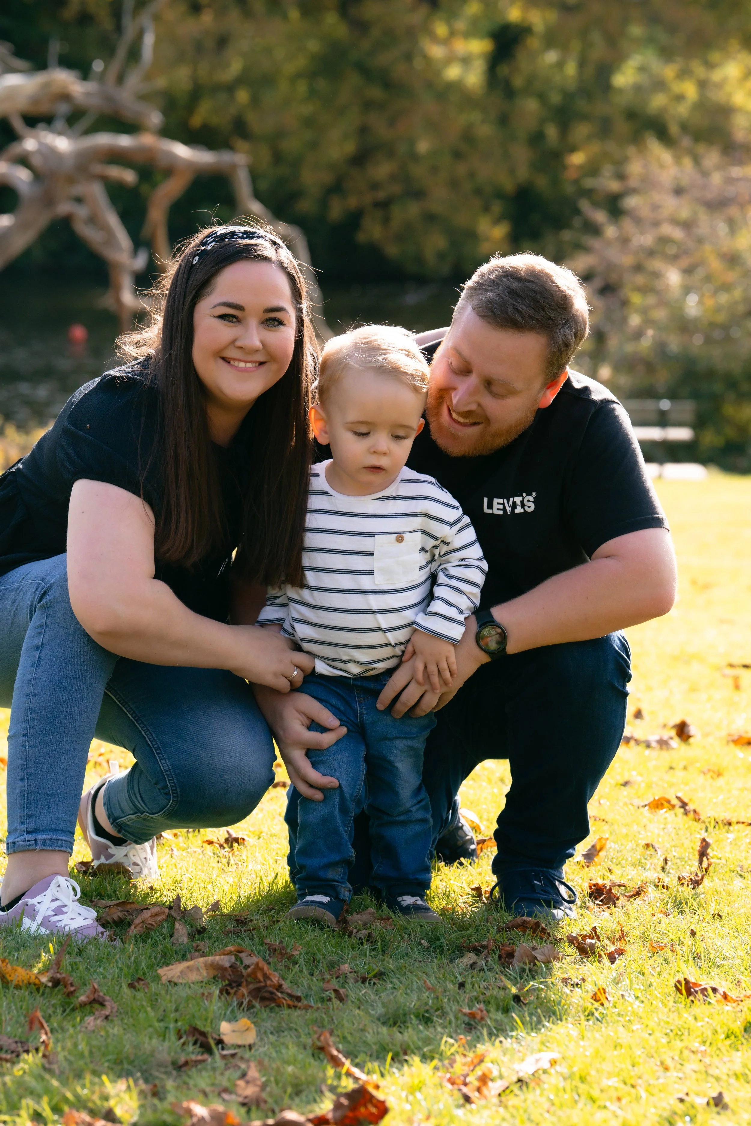 A happy family of three outdoors in a park on a sunny autumn day, with leaves scattered on the grass.