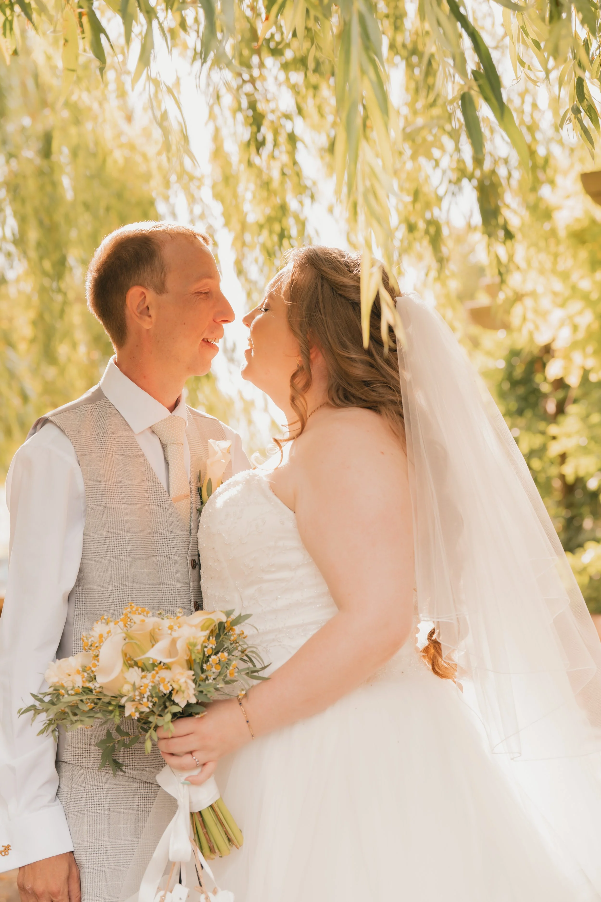 Two women smiling and looking at each other, one holding a bouquet of white flowers, the other wearing a white off-the-shoulder dress with tattoos visible on her shoulders and chest.