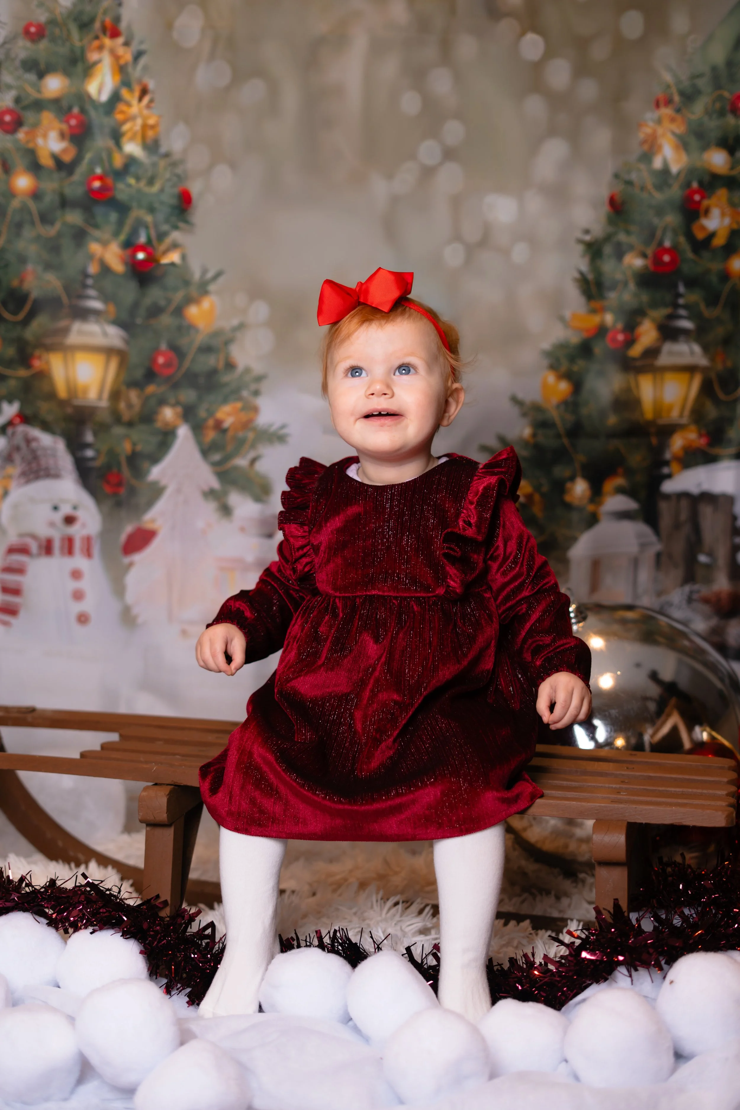 A young girl wearing a red velvet dress and a red bow in her hair, sitting on a wooden bench surrounded by white cotton balls, with Christmas trees and holiday decorations in the background.