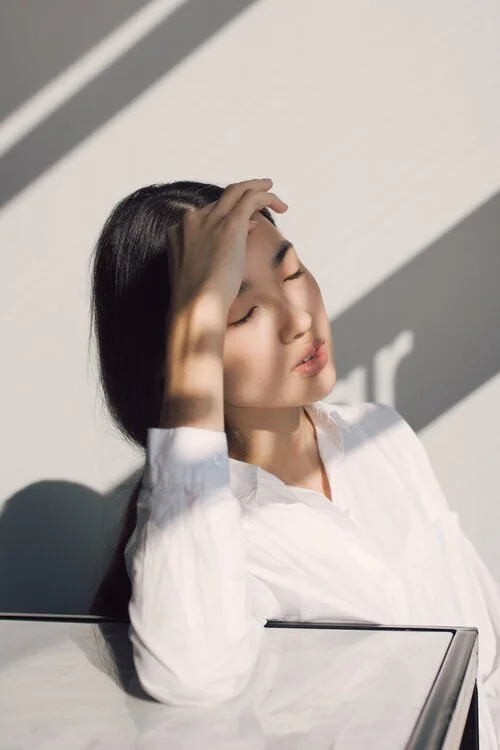 A woman with long dark hair and a white shirt resting her head and hand on her forehead with closed eyes, sitting beside a mirror with sunlight casting shadows on a white wall.