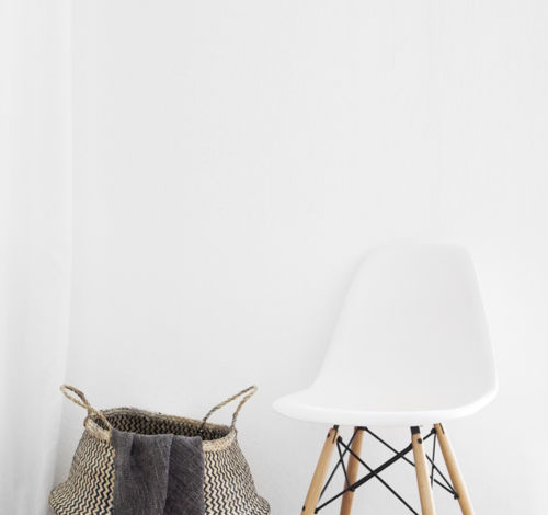 Minimalist white chair with wooden legs and black supports next to a woven basket with fabric in a white room.