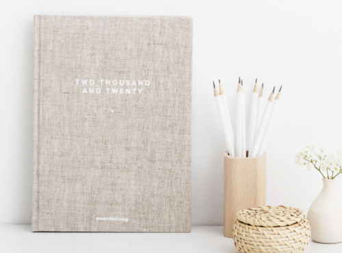 A beige notebook with white text reading 'TWO THOUSAND AND TWENTY' on a white surface, next to a wooden holder with white pens, a small woven basket, and a white vase with a small white flower.