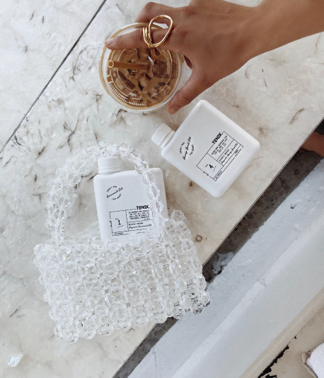 A hand holding a glass filled with iced coffee, next to two bottles of liquid soap or sanitizer on a marble surface, with one bottle surrounded by soap bubbles.