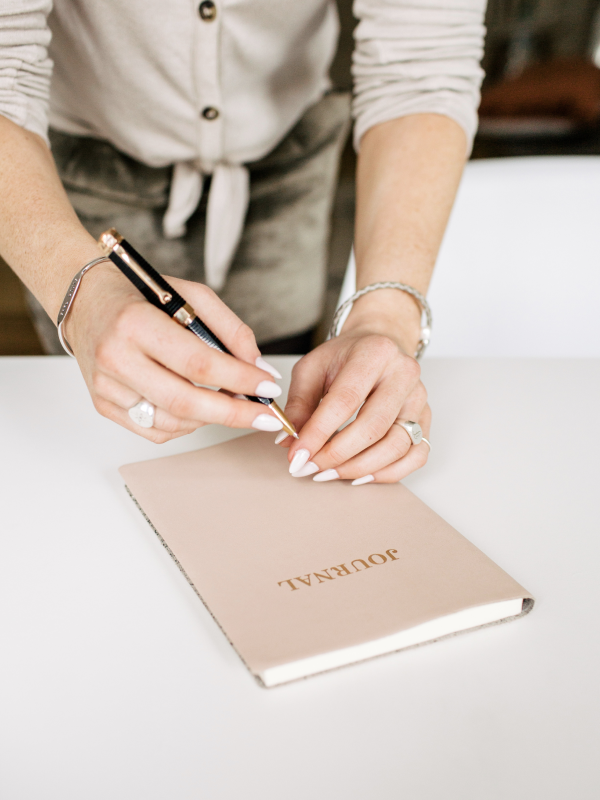 A person signing a beige journal titled "JOURNAL" on a white table, wearing a white button-up shirt and jewelry.