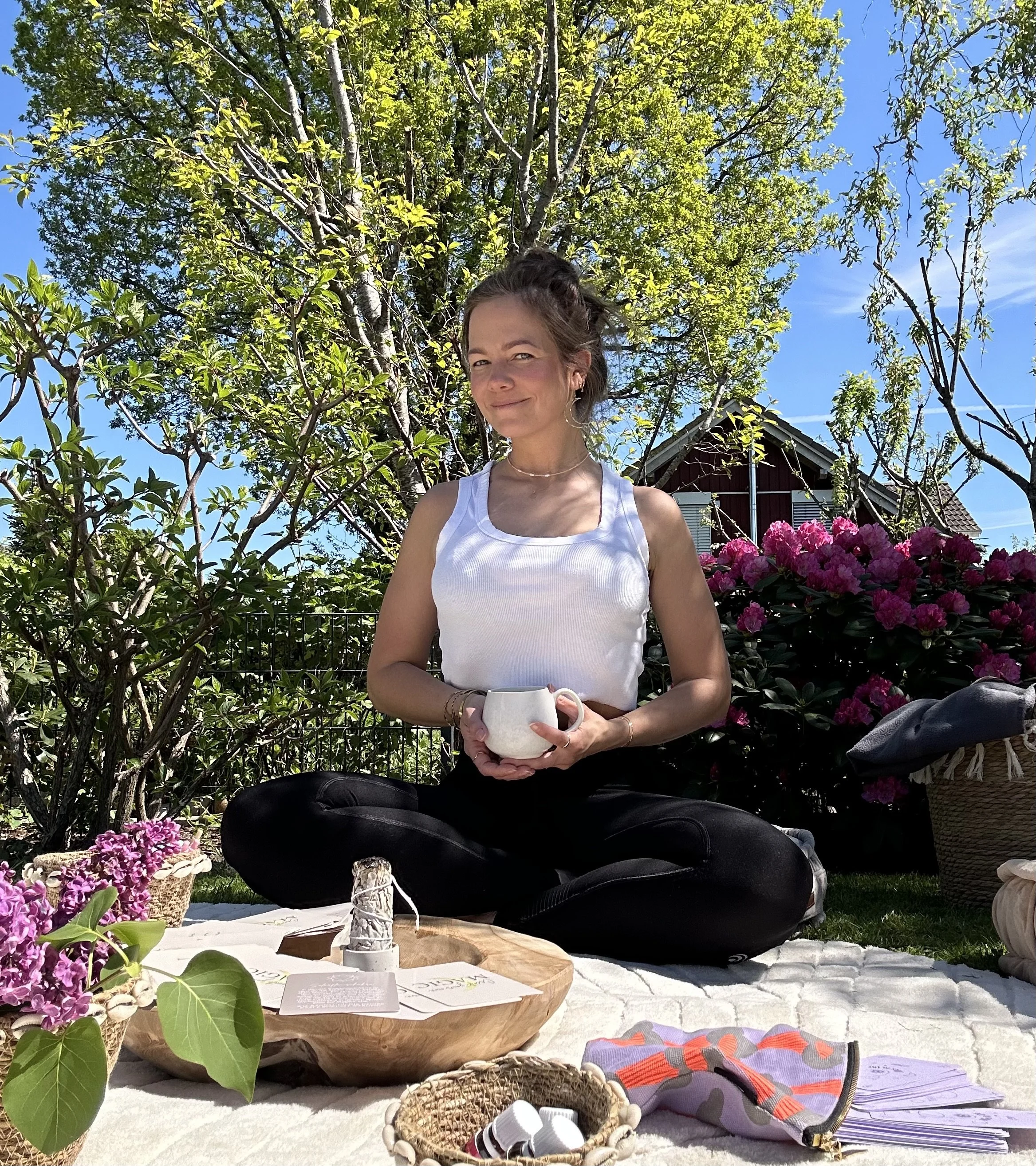 Junge Frau sitzt im Garten auf einem weißen Tuch, hält eine weiße Tasse, umgeben von Blumen, Bäumen und einem blauen Himmel, mit einer Holschale, Papiersorten, einer kleinen Tasche und anderen Gegenständen im Vordergrund.