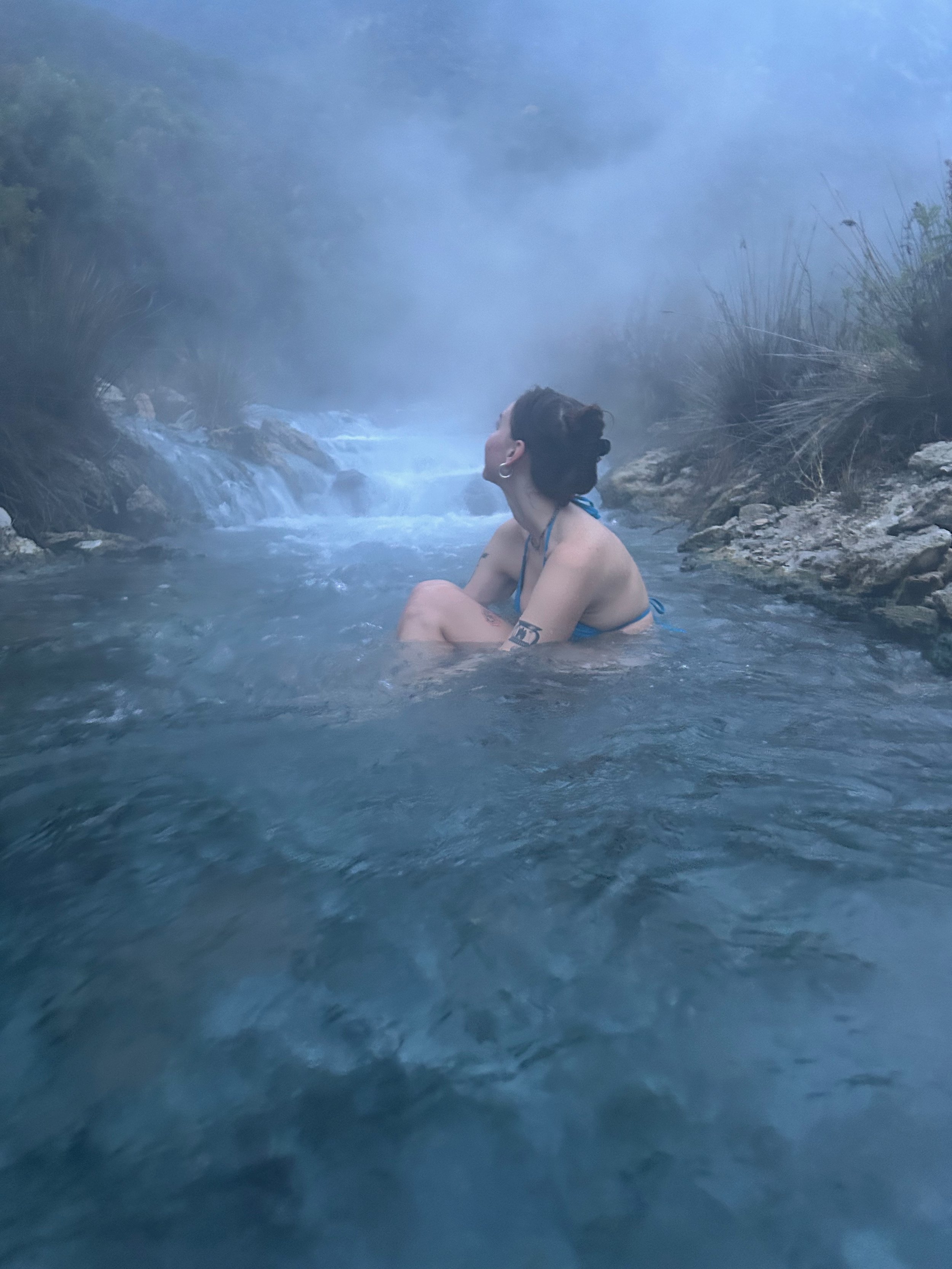 A woman sitting in a hot spring or steam bath, surrounded by rocks and plants, with mist or steam rising around her.