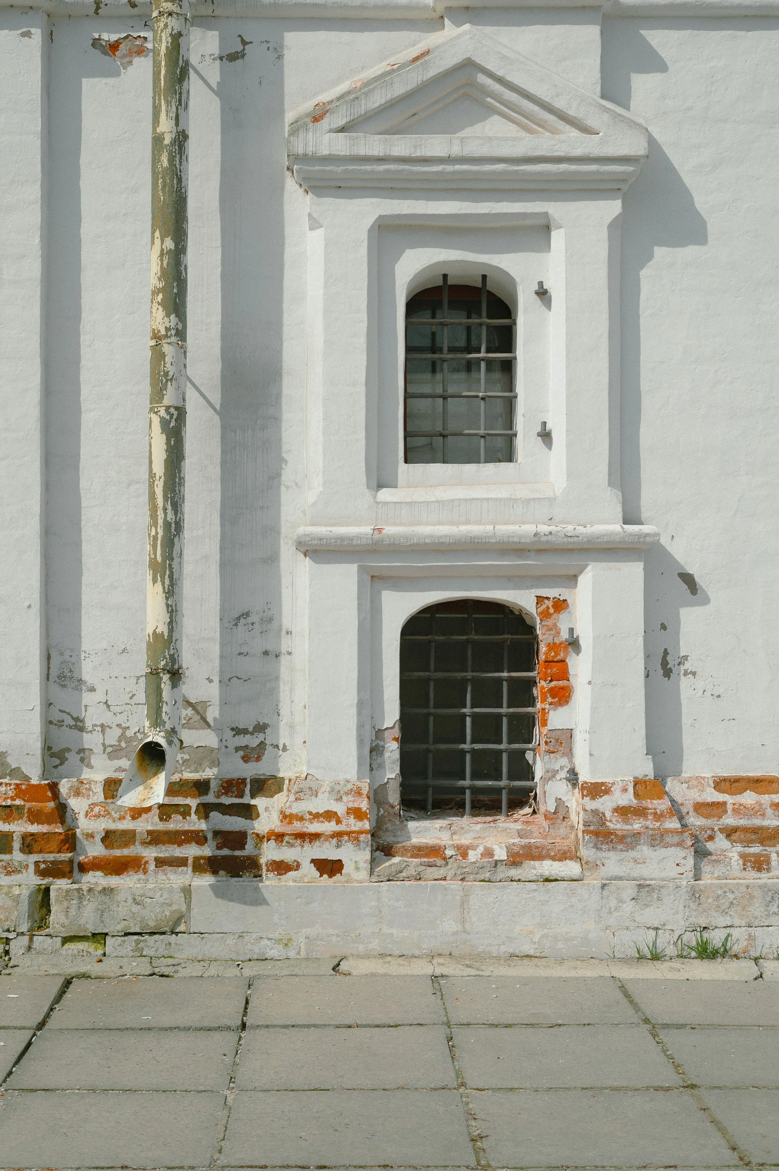 A white building with brick accents and a gray drainpipe. Two small barred windows, one above the other, with some damage to the wall and bricks exposed.