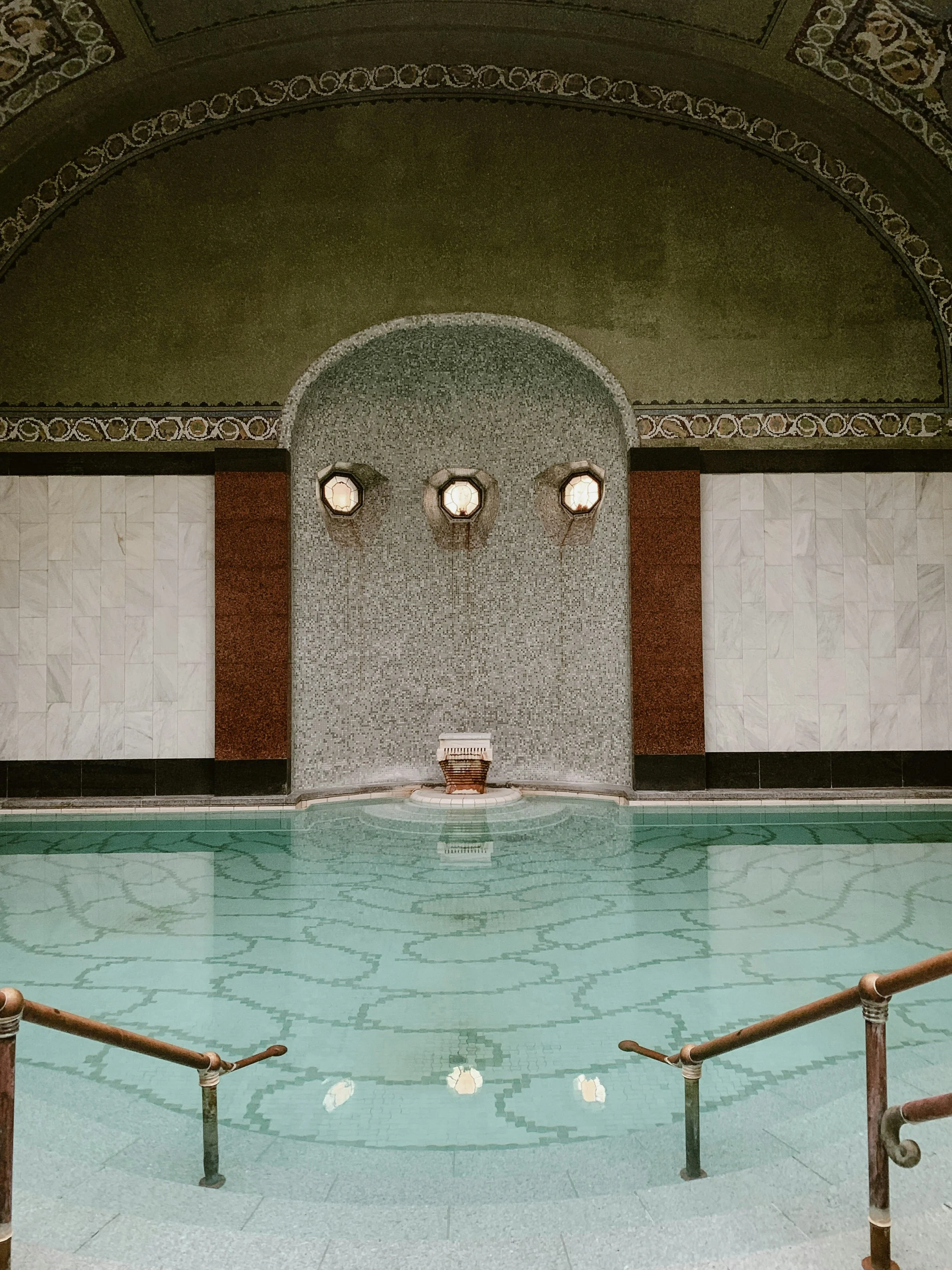 Indoor vintage swimming pool with ornate ceiling, decorative wall, and water feature with three small lights above.