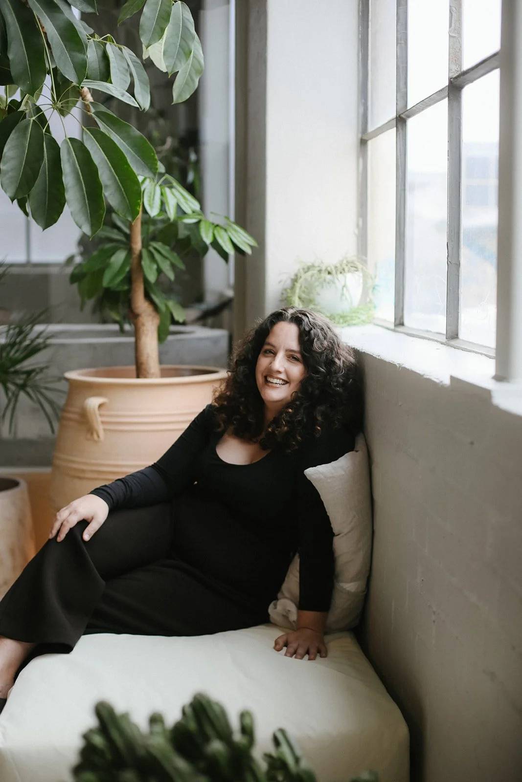 A woman with curly black hair smiling and lounging on a bed or sofa near a large window, with green potted plants in the background.