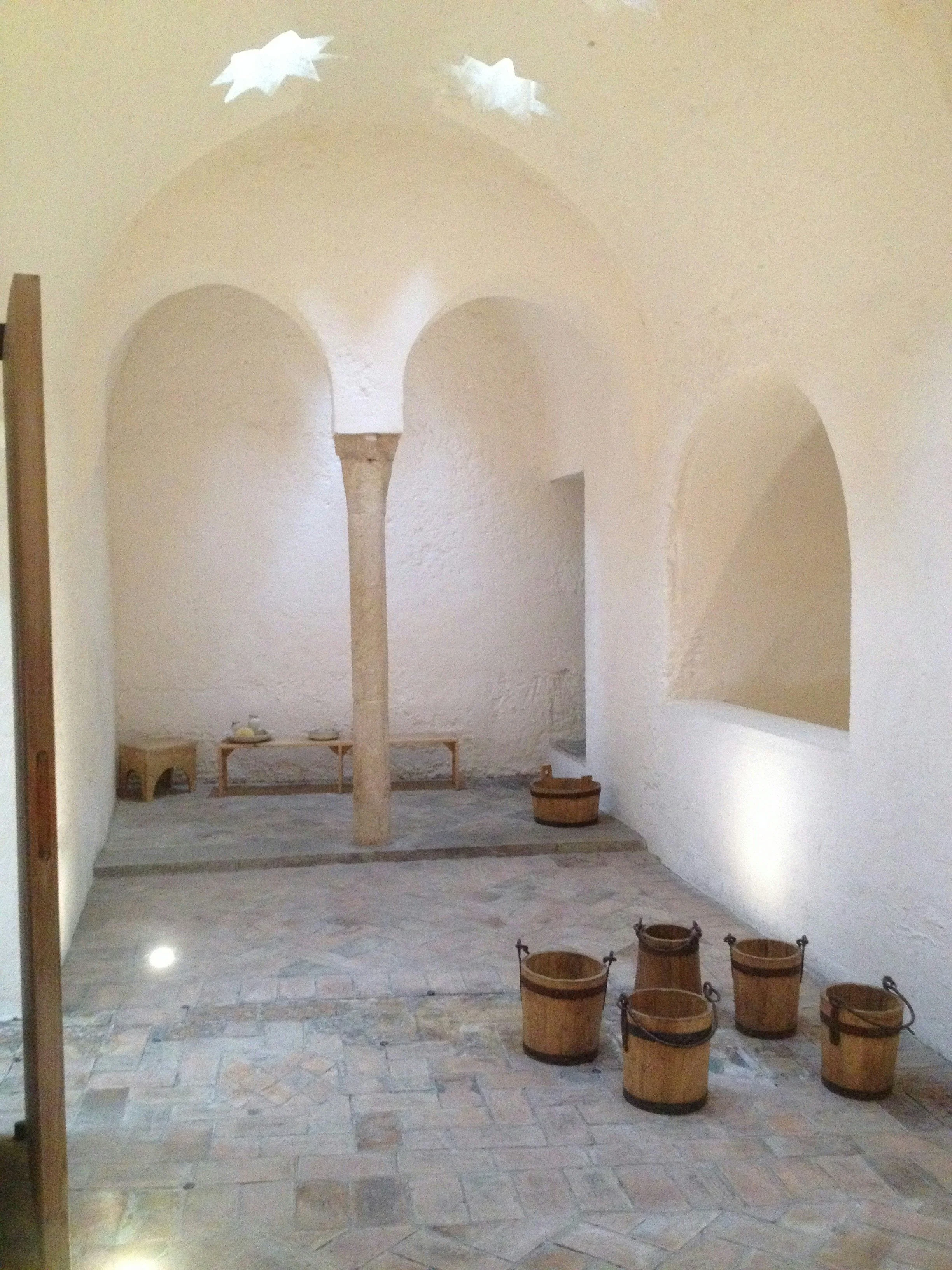 Interior of a room with white textured walls, arched openings, and a stone central column. Wooden buckets are on the floor, with a small wooden table holding a few items in the back.