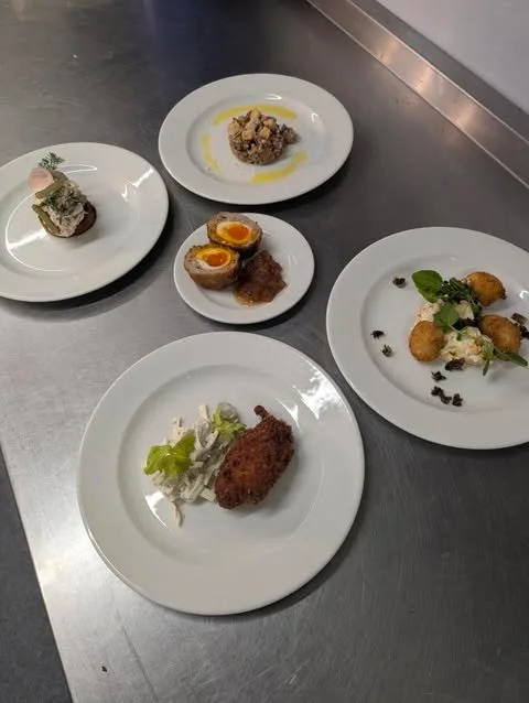 Assorted gourmet dishes on white plates including an appetizer, an egg-centered dish, deep-fried food, and a salad with greens on a stainless steel counter.