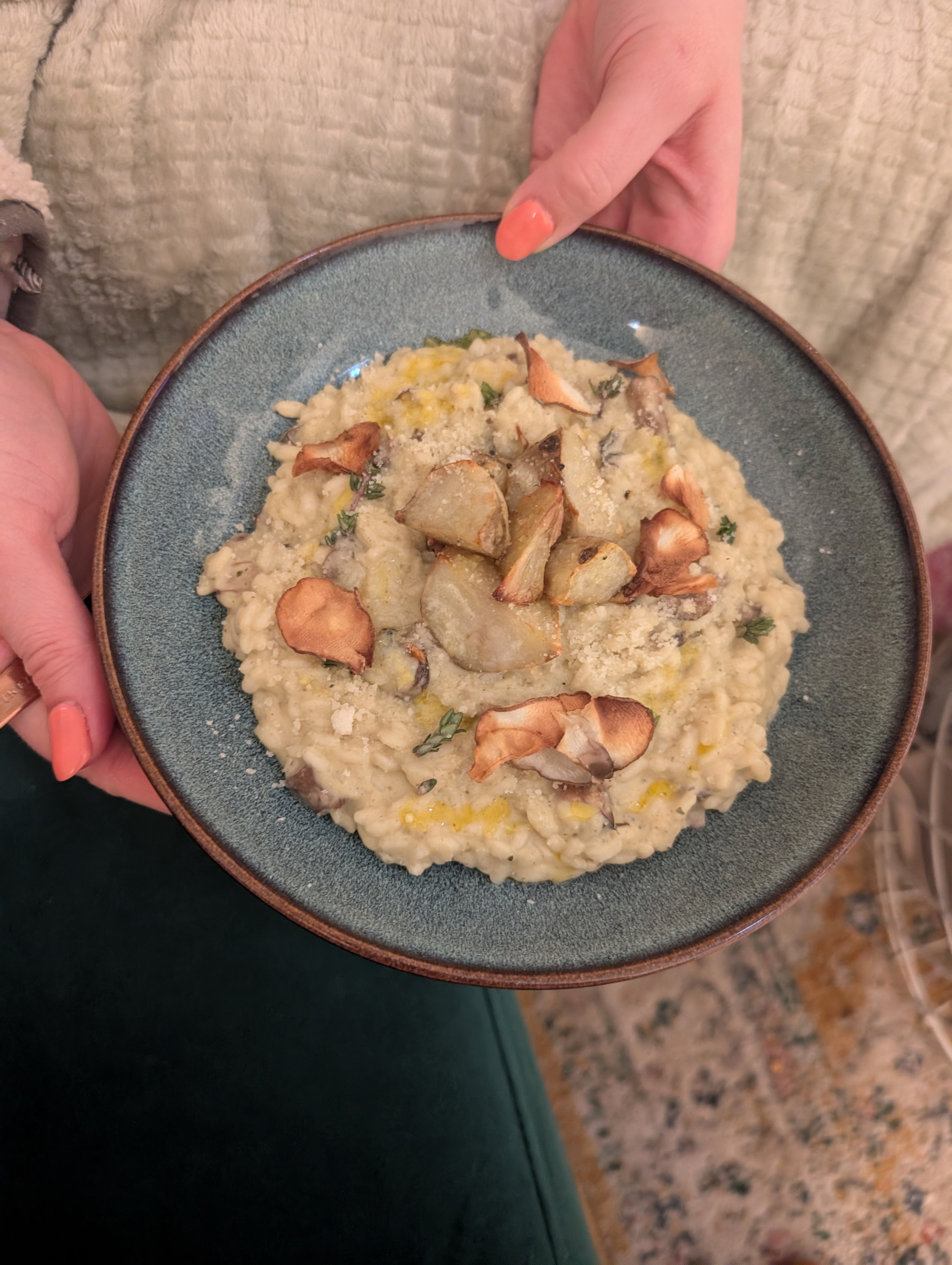 A person holding a blue ceramic bowl of creamy risotto topped with roasted garlic and herbs.