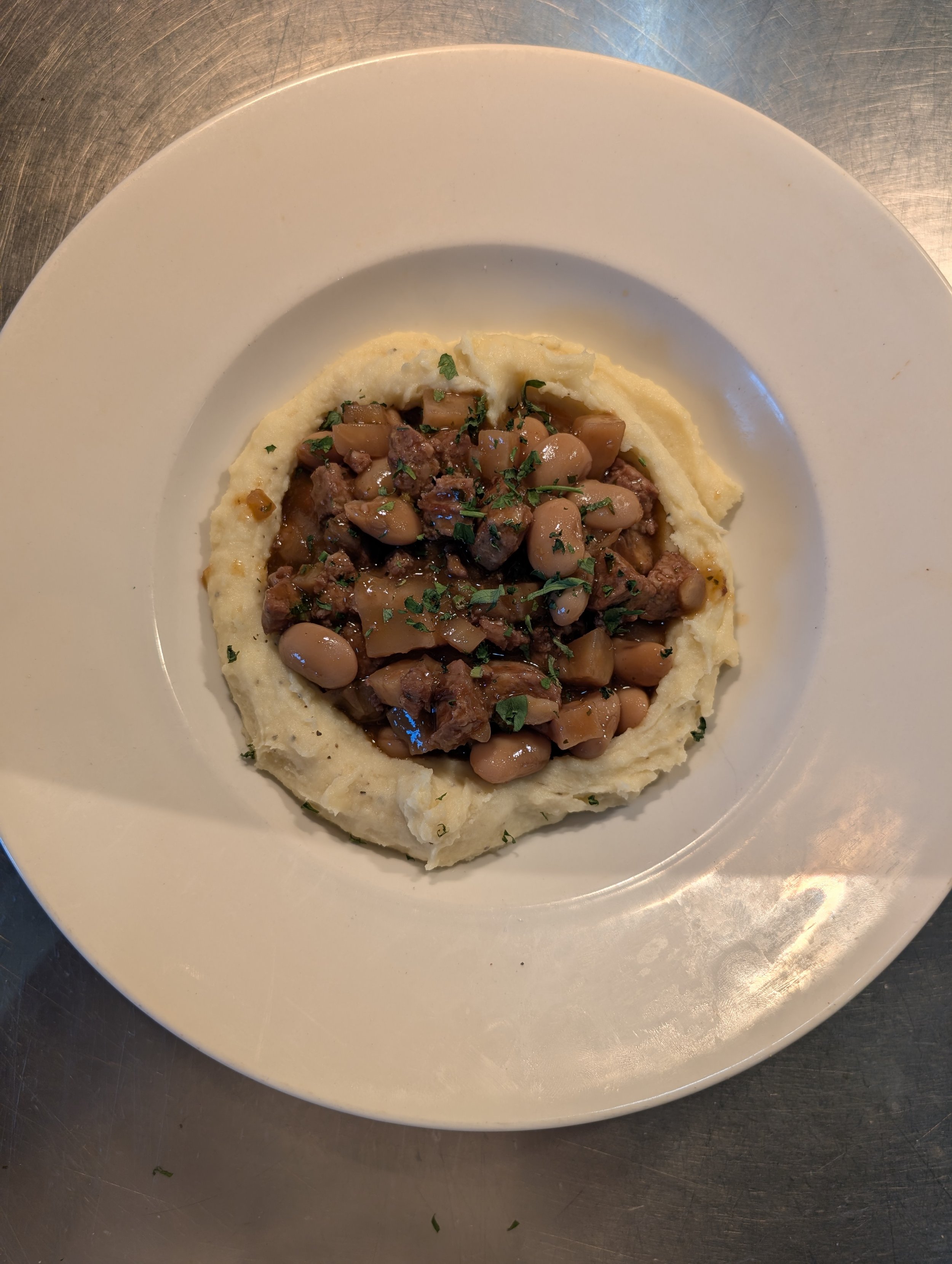 A white plate with mashed potatoes topped with a stew of beans, beef, and chopped herbs.