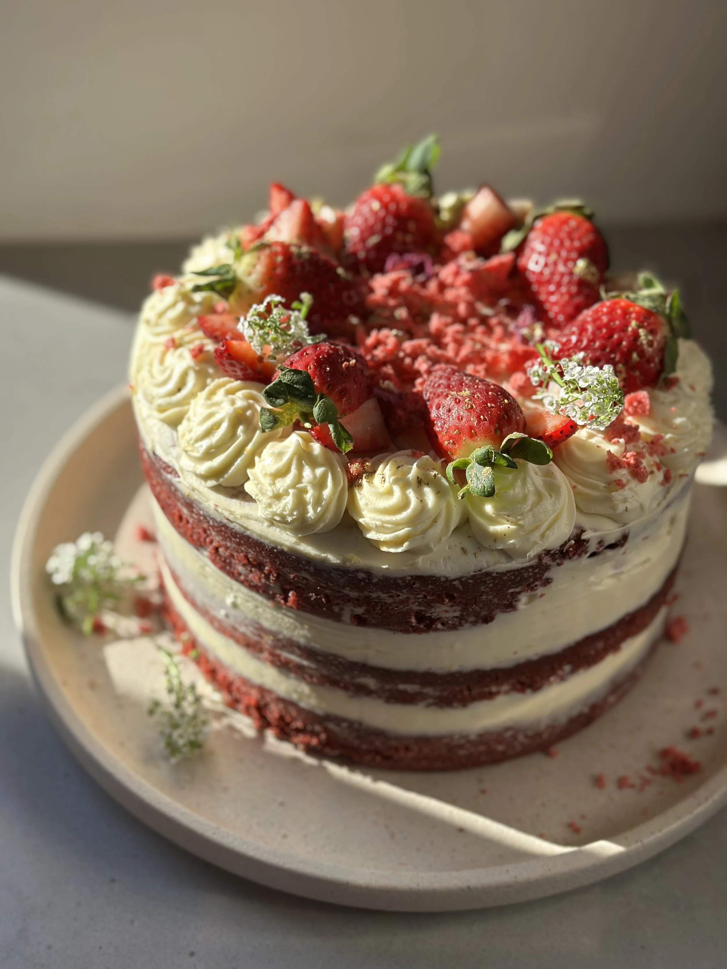 A layered red velvet cake with white cream frosting, topped with fresh strawberries, small white flowers, and red sprinkles, on a round beige plate.