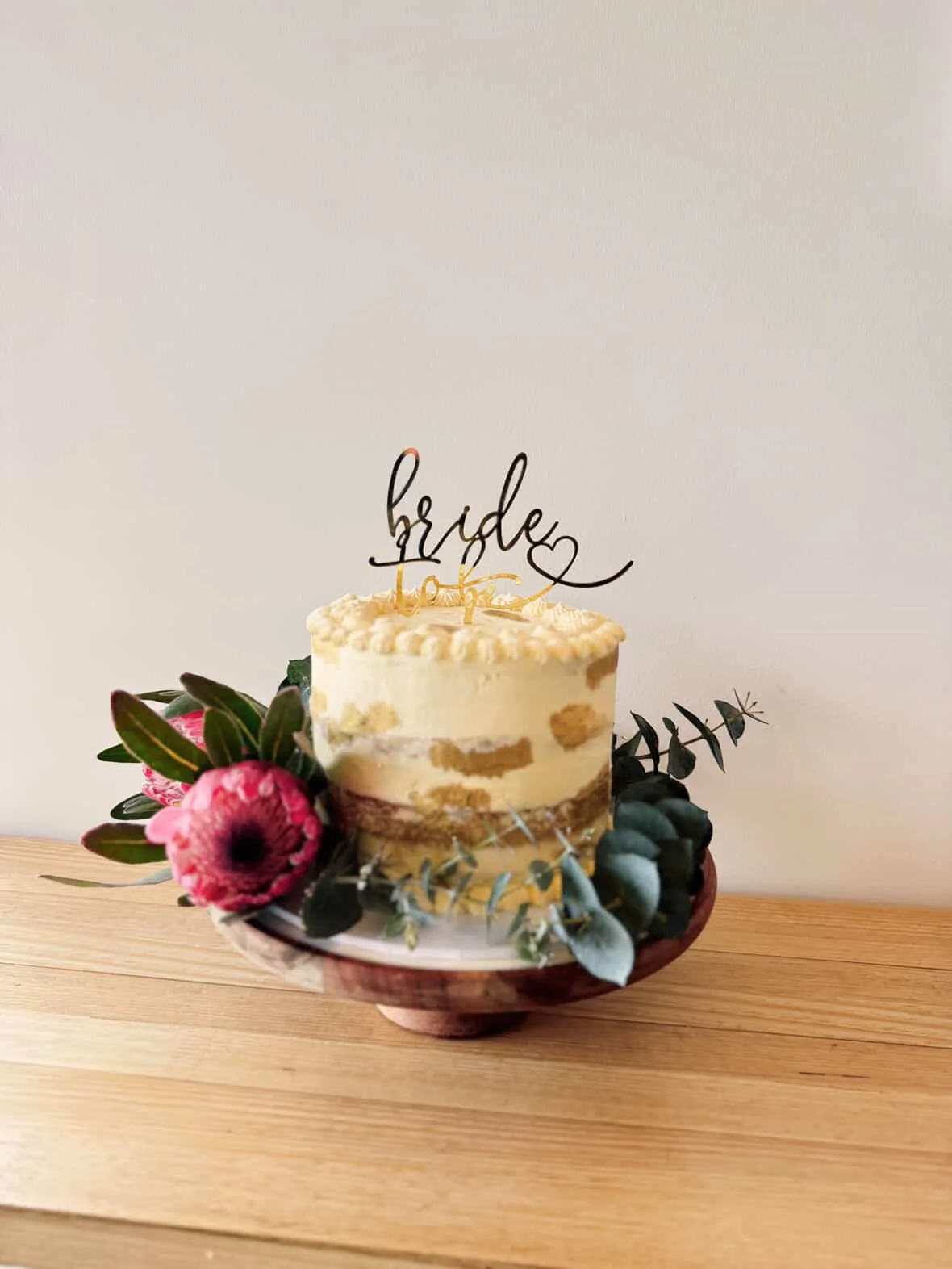 A decorated wedding cake with a 'bride' topper, surrounded by flowers and greenery, on a wooden table.