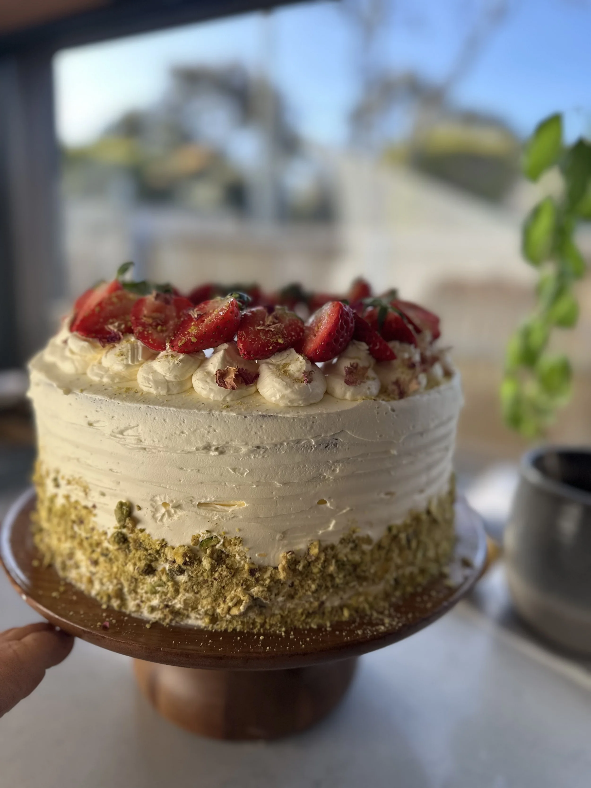A round layered cake with white frosting, topped with sliced strawberries and decorative cream, with a crumbly pistachio or nut coating around the sides, placed on a brown cake stand.