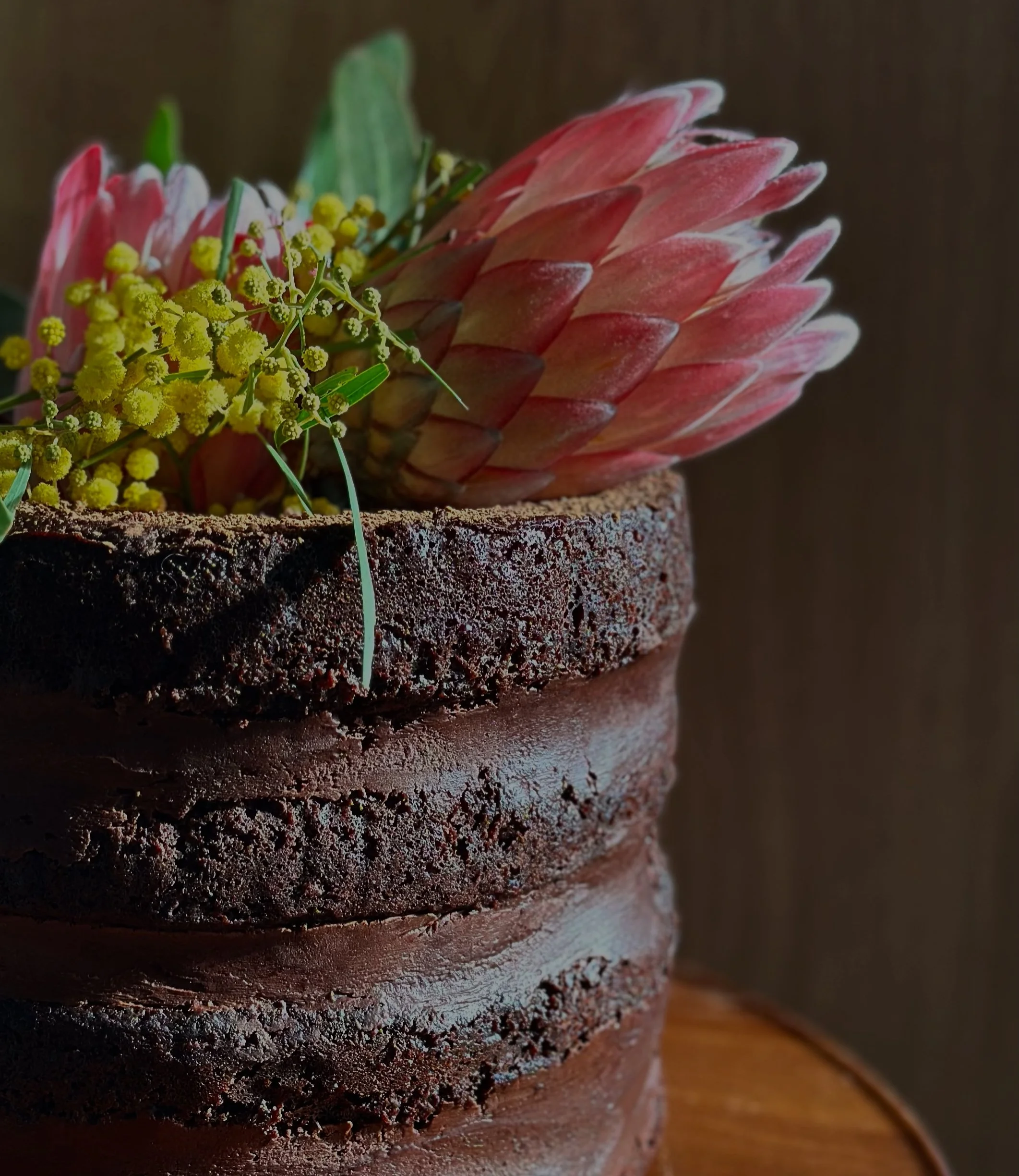 Chocolate layered cake decorated with pink protea flower, yellow sprigs, and green leaves.