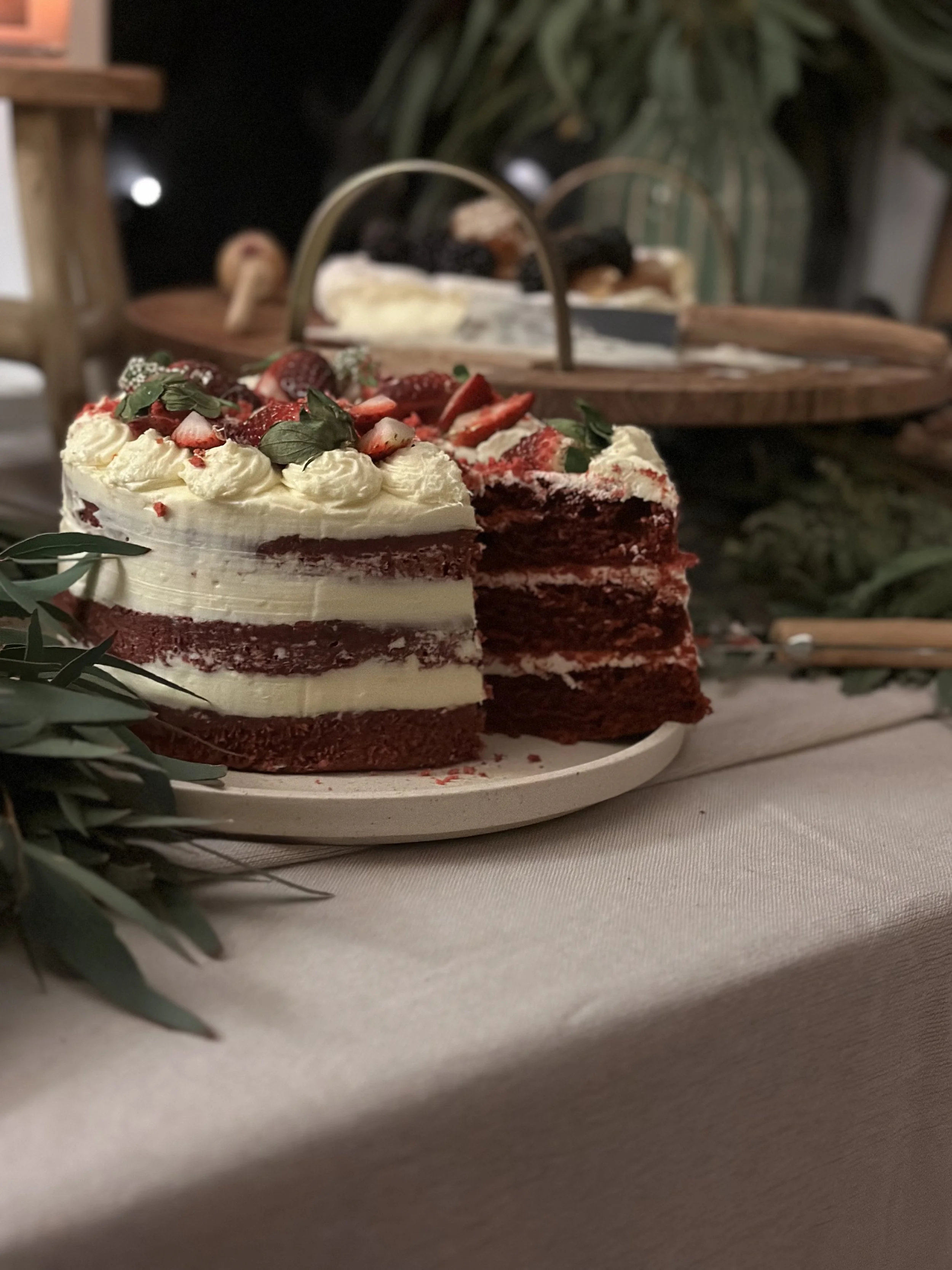 A layered red velvet cake with white cream cheese frosting topped with strawberries and greenery, placed on a white plate on a table with a beige tablecloth.