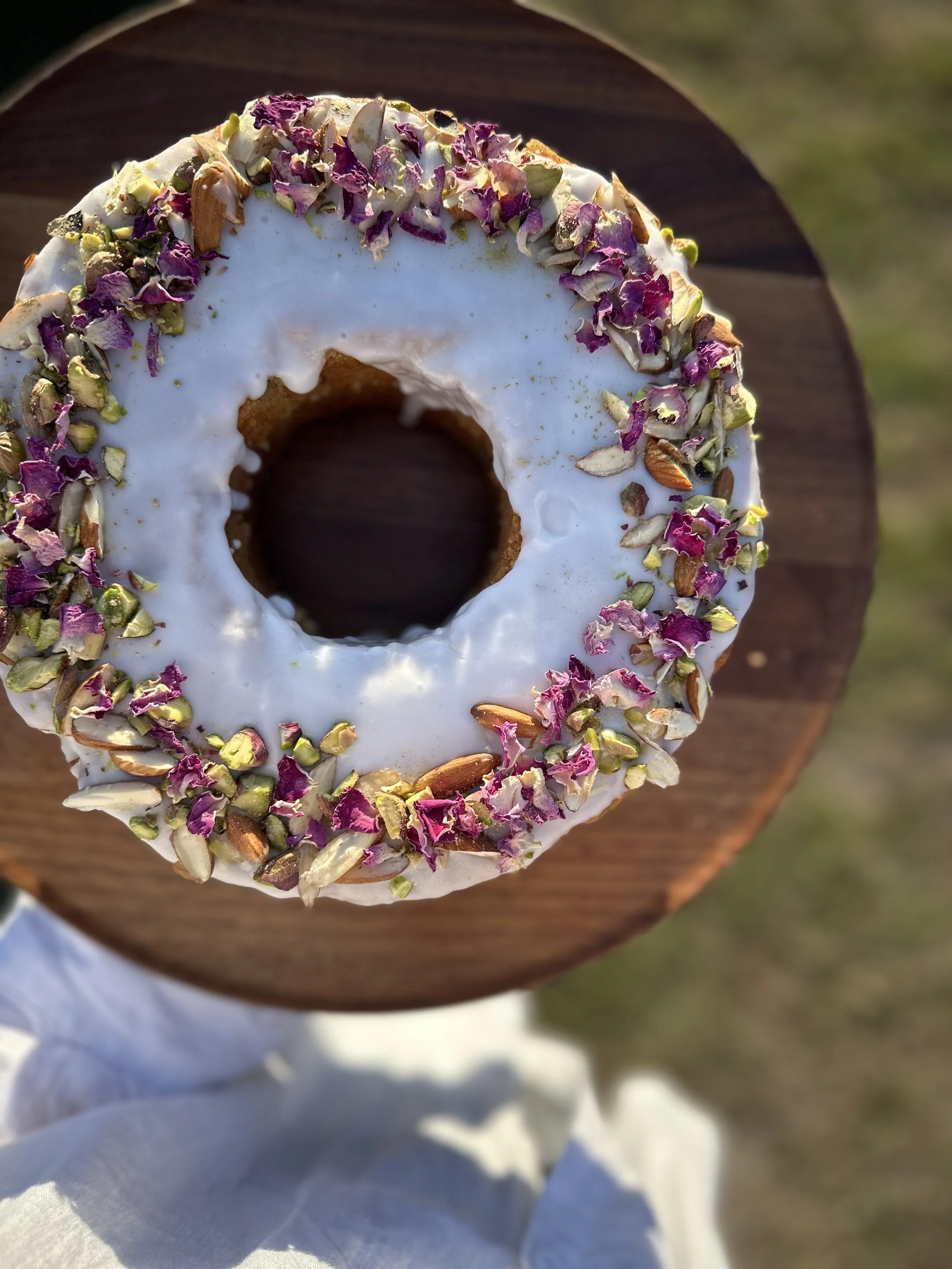 Top-down view of a donut with white glaze and sprinkled with dried rose petals and nuts, placed on a wooden surface.