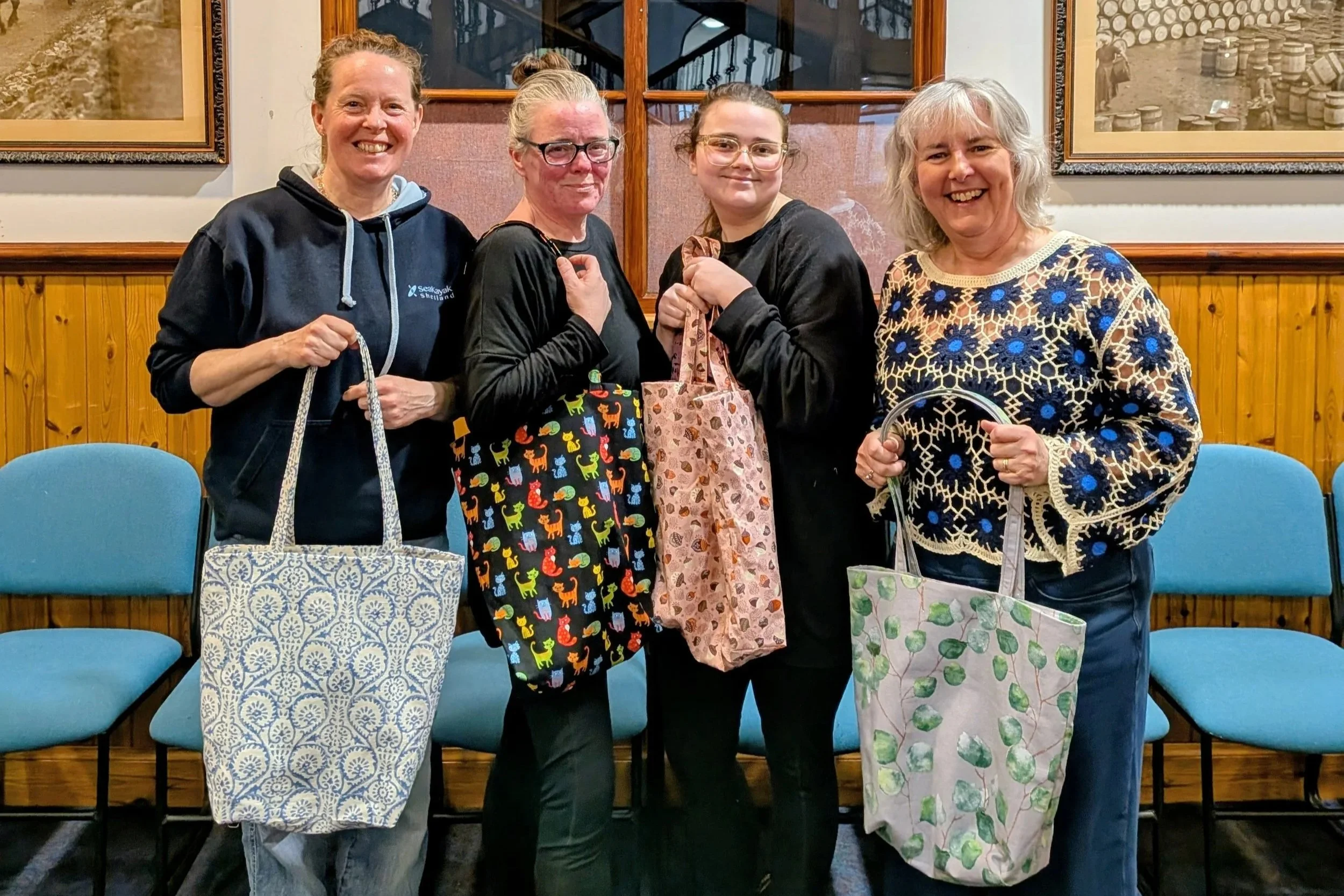 Four women pose holding handmade tote bags. They stand in a line and smile at the camera