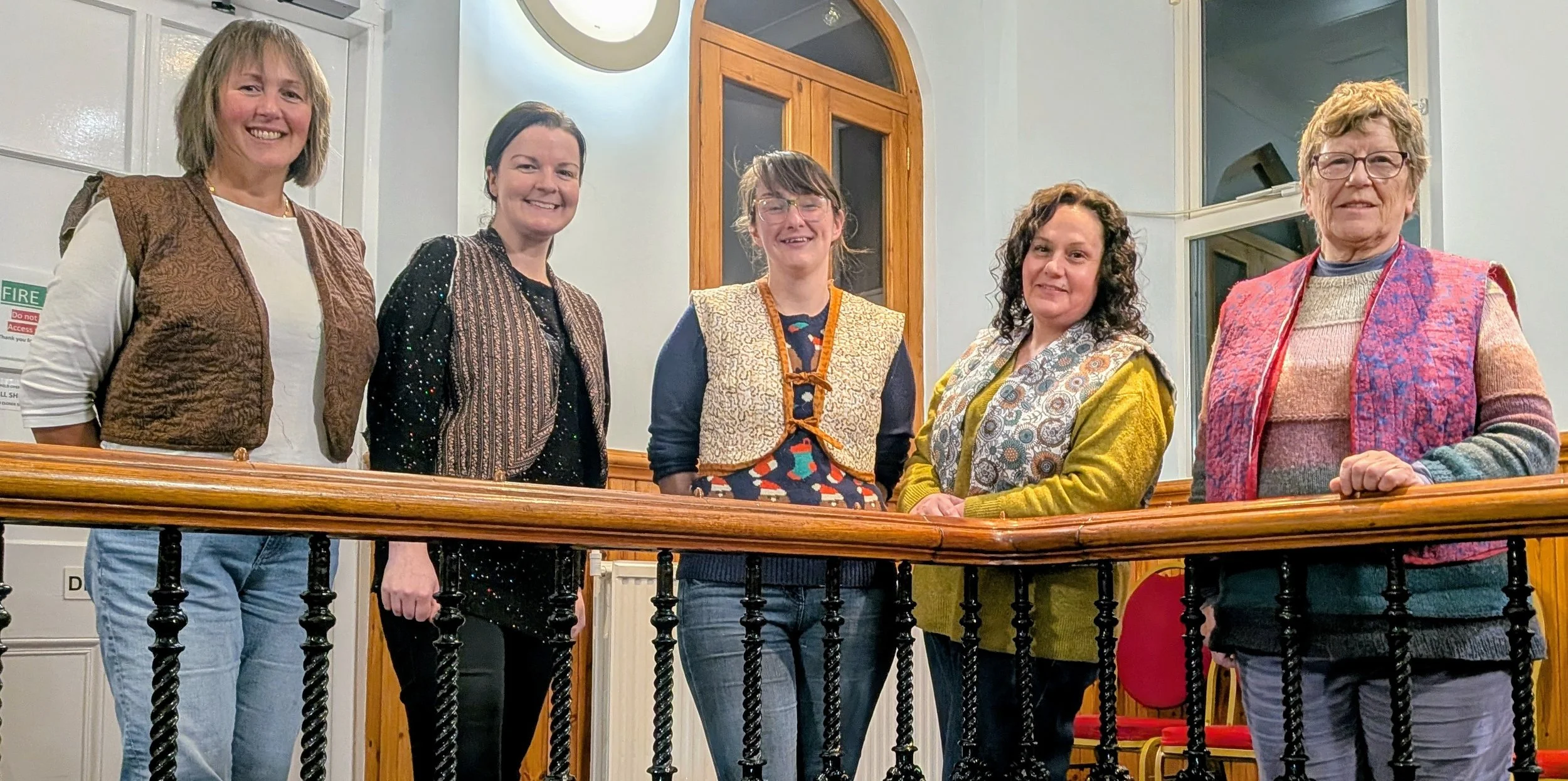 Five women lean on a handrail wearing quilted waistcoats in a variety of fabrics and colours.