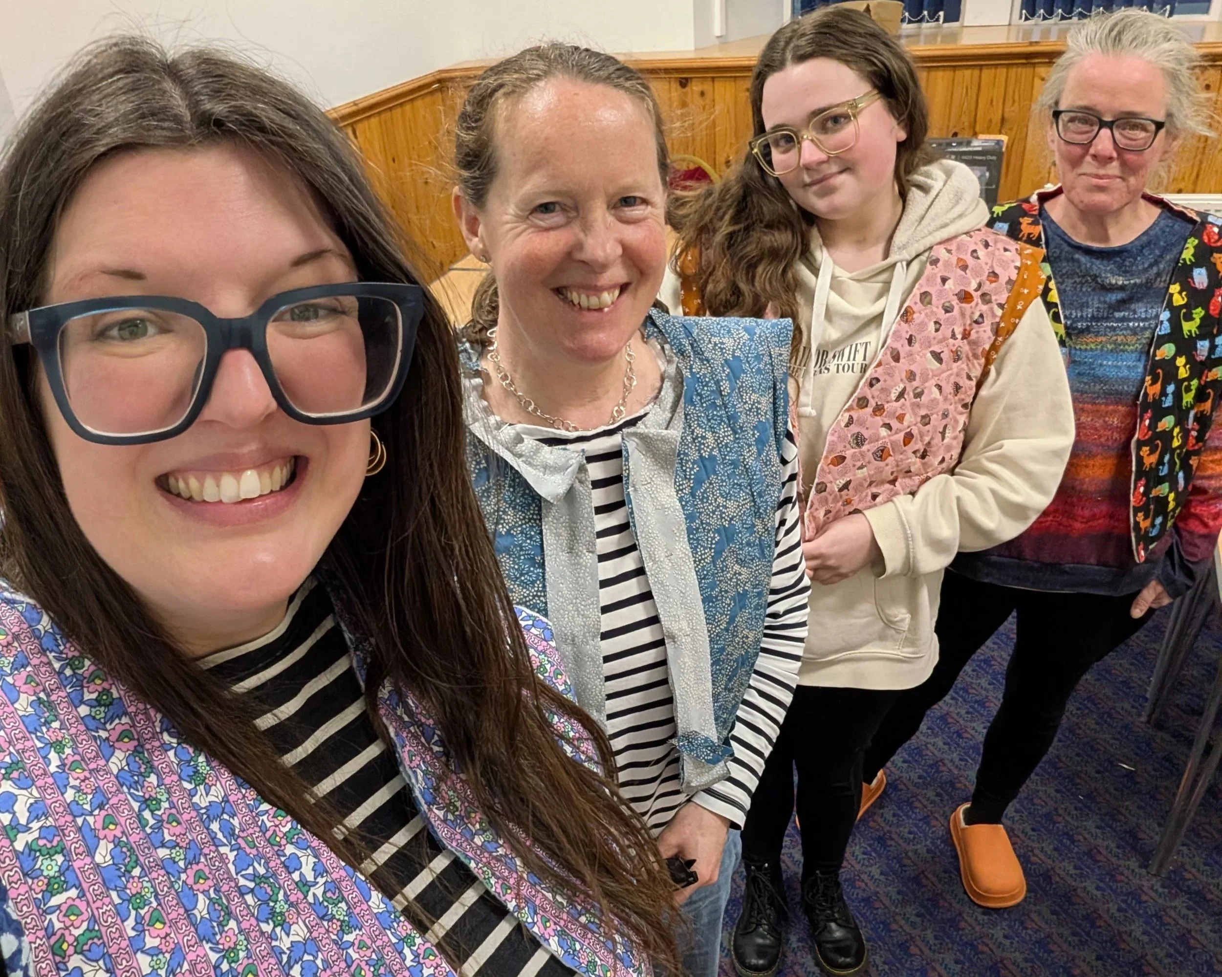 Four smiling women pose for a selfie wearing almost-finished quilted waistcoats