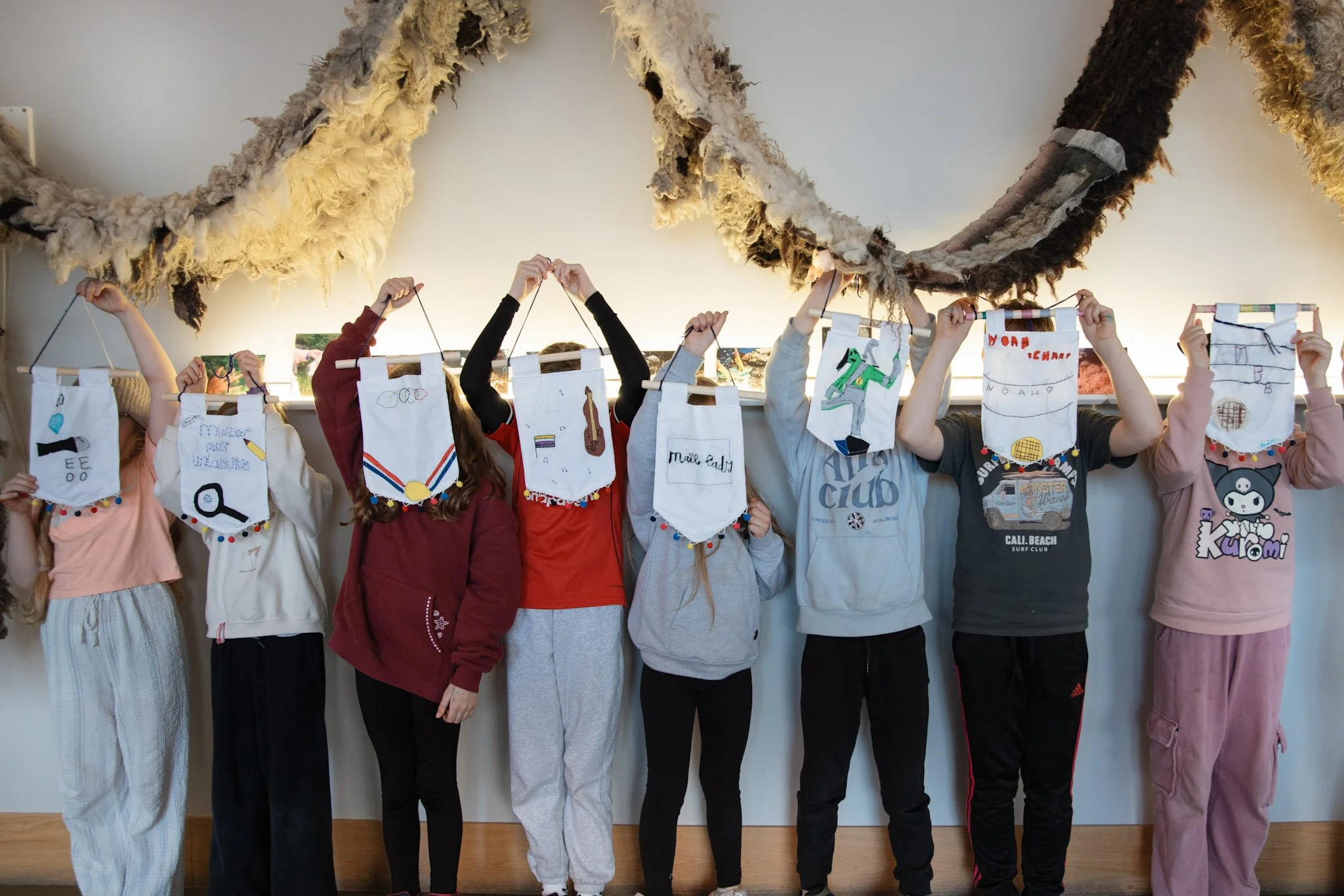 In the foyer at Mareel, eight children stand in a row holding small handmade banners in front of their faces. The banners are embroidered and appliqued and finished with a pompom trim at the bottom.
