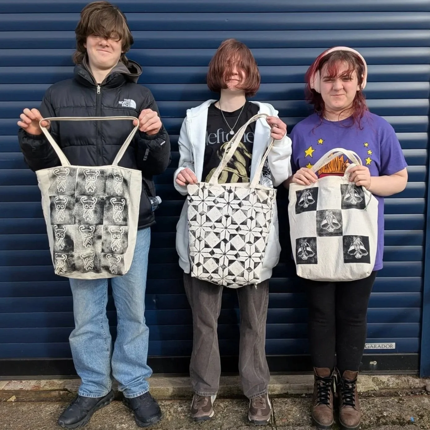 Three teenagers stand in front of navy blue shutters, holding black and white printed tote bags.