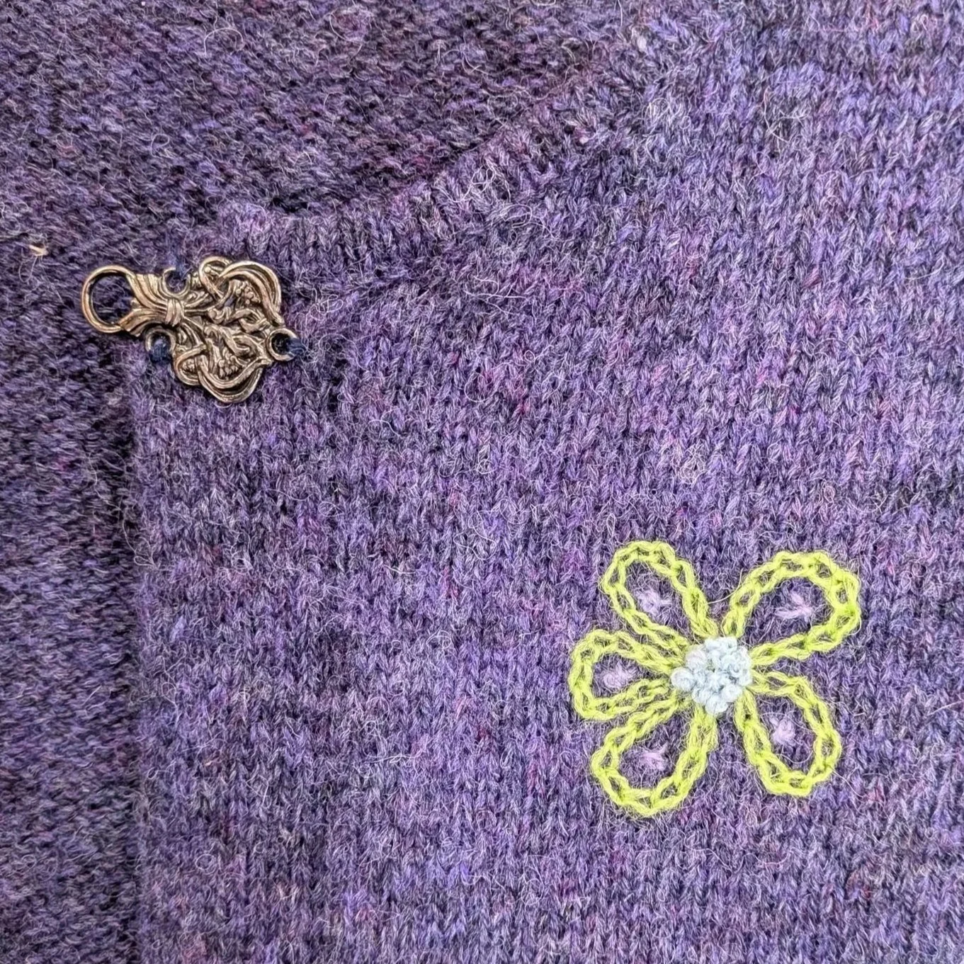 A small green and blue flower, made with chain stitch and French knots, is embroidered onto a purple wool cardigan with an ornate silver clasp.