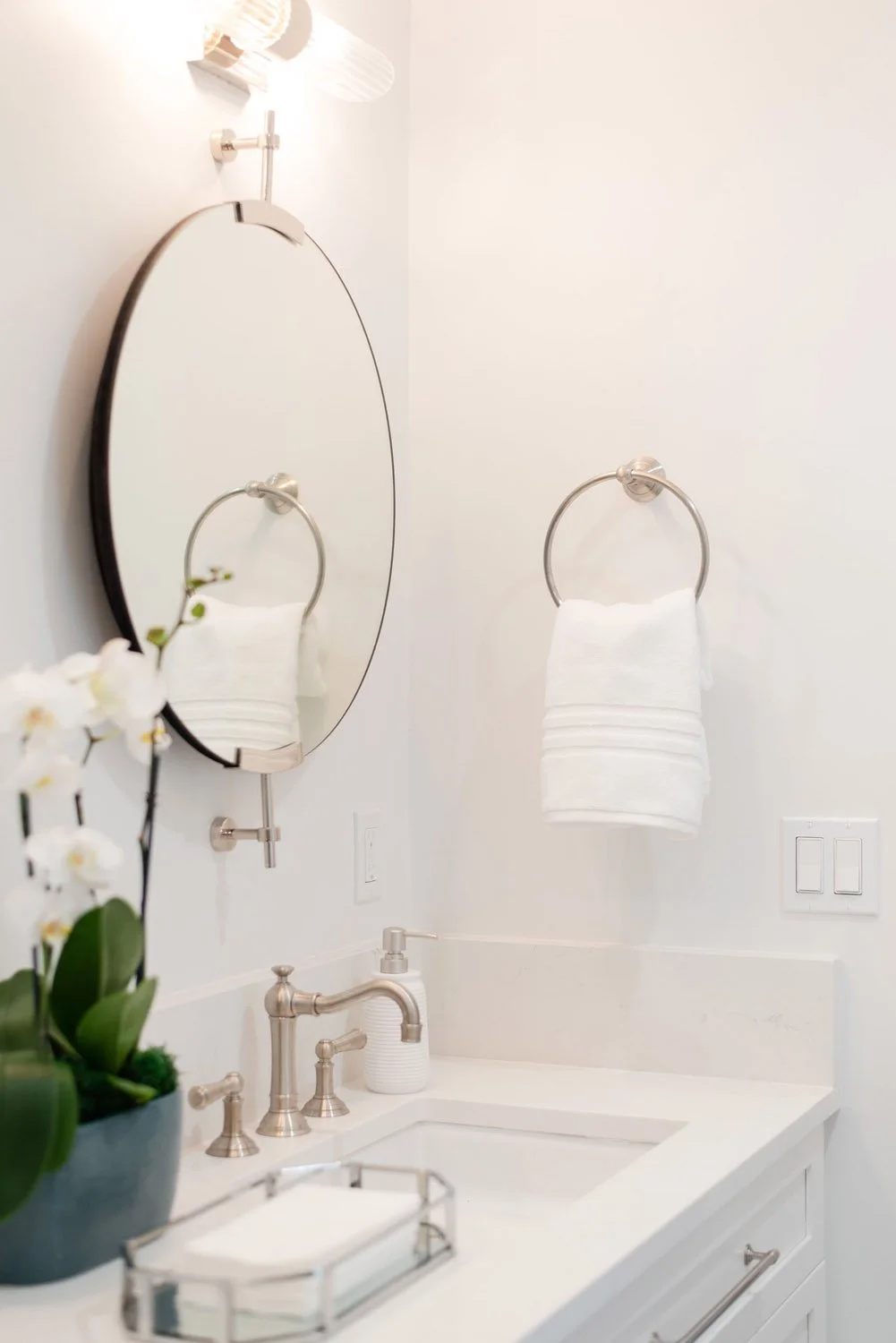Round mirror and towel ring above a white vanity with orchid