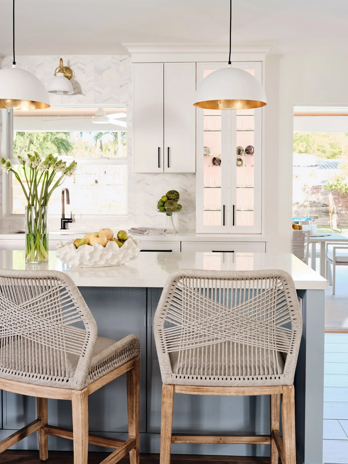 Woven counter stools at a white kitchen island with brass pendants