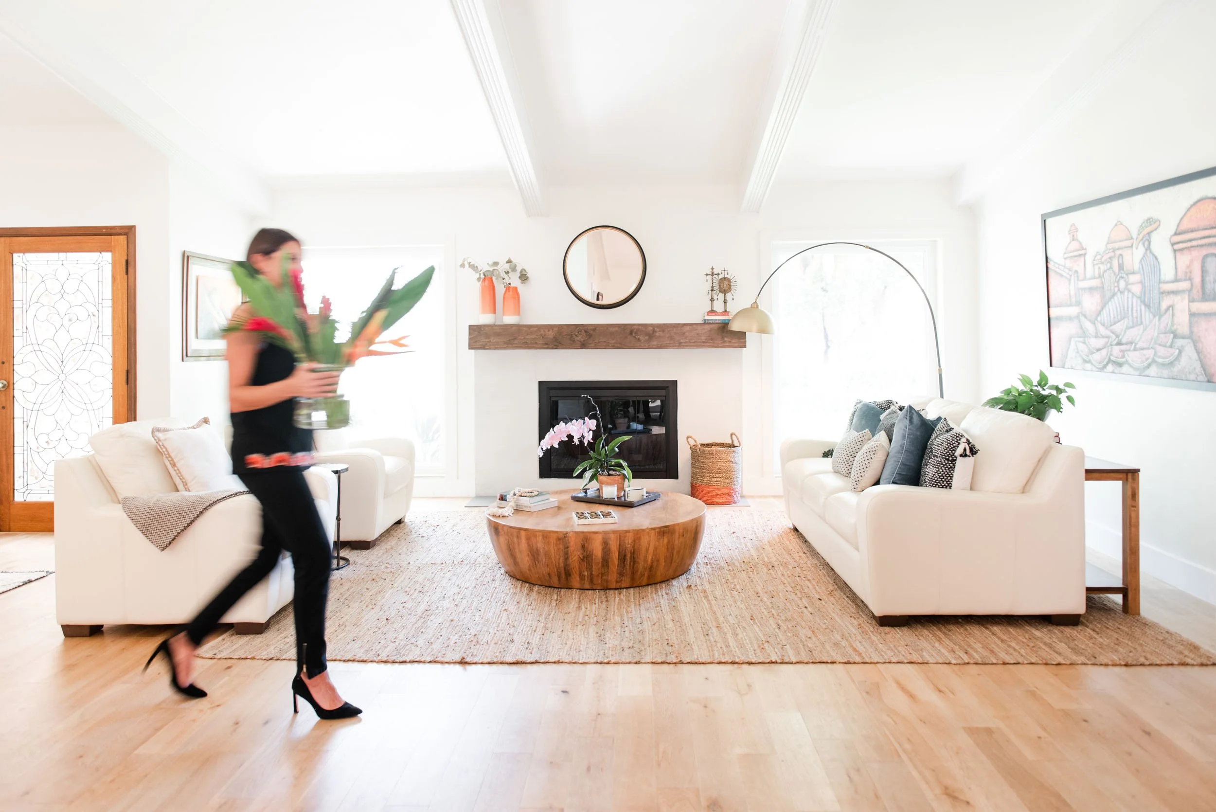 Bright, modern living room with white sofas, a round wooden coffee table, fireplace, and a person carrying flowers.