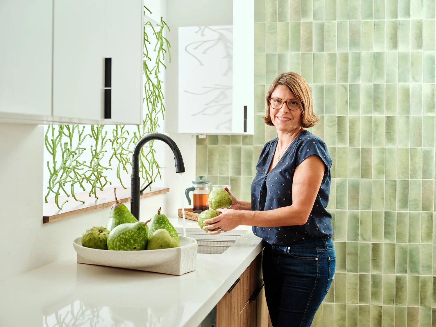 Woman holding a bowl of pears at a kitchen sink with green tile backsplash