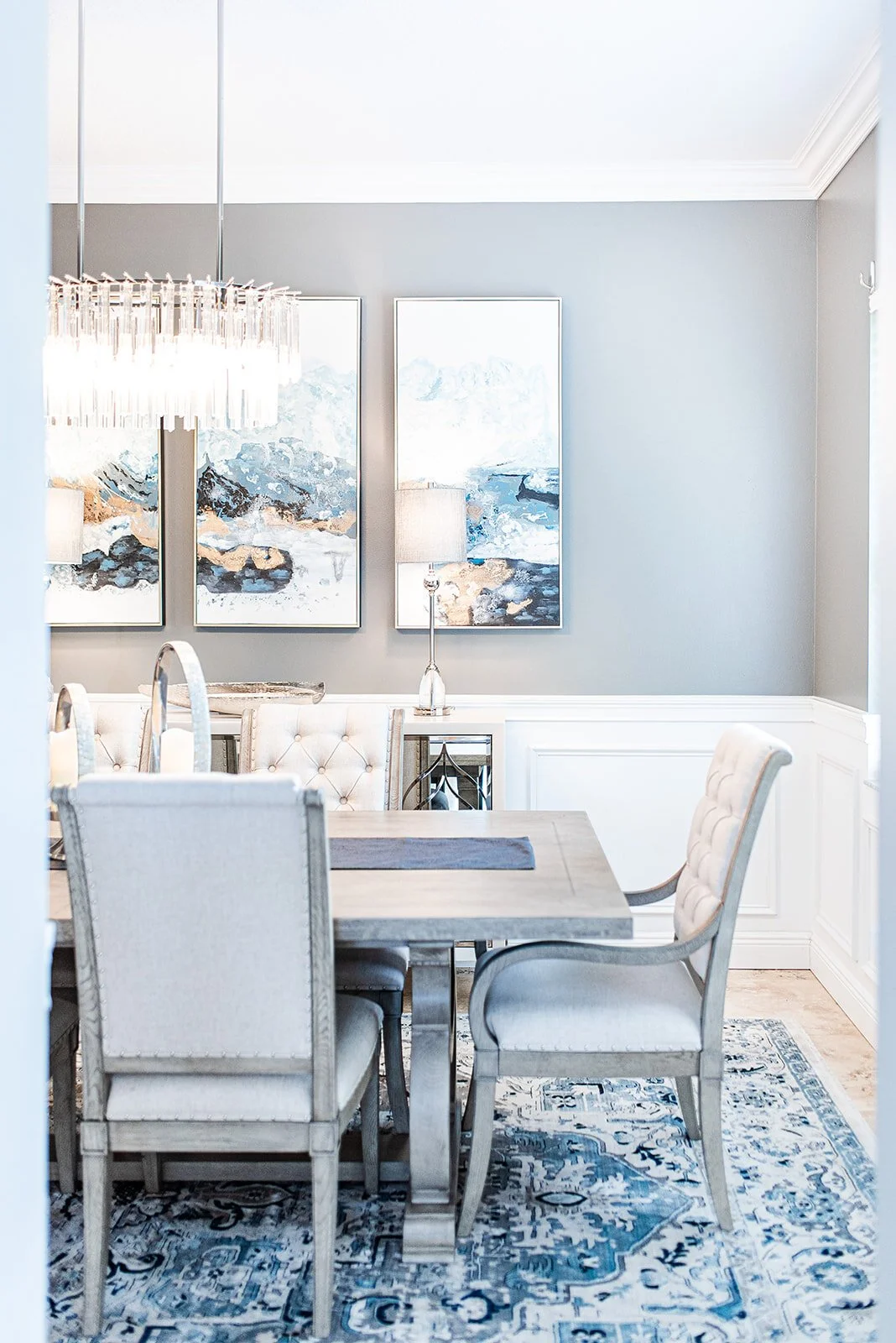 Dining table with gray chairs, chandelier and three paintings