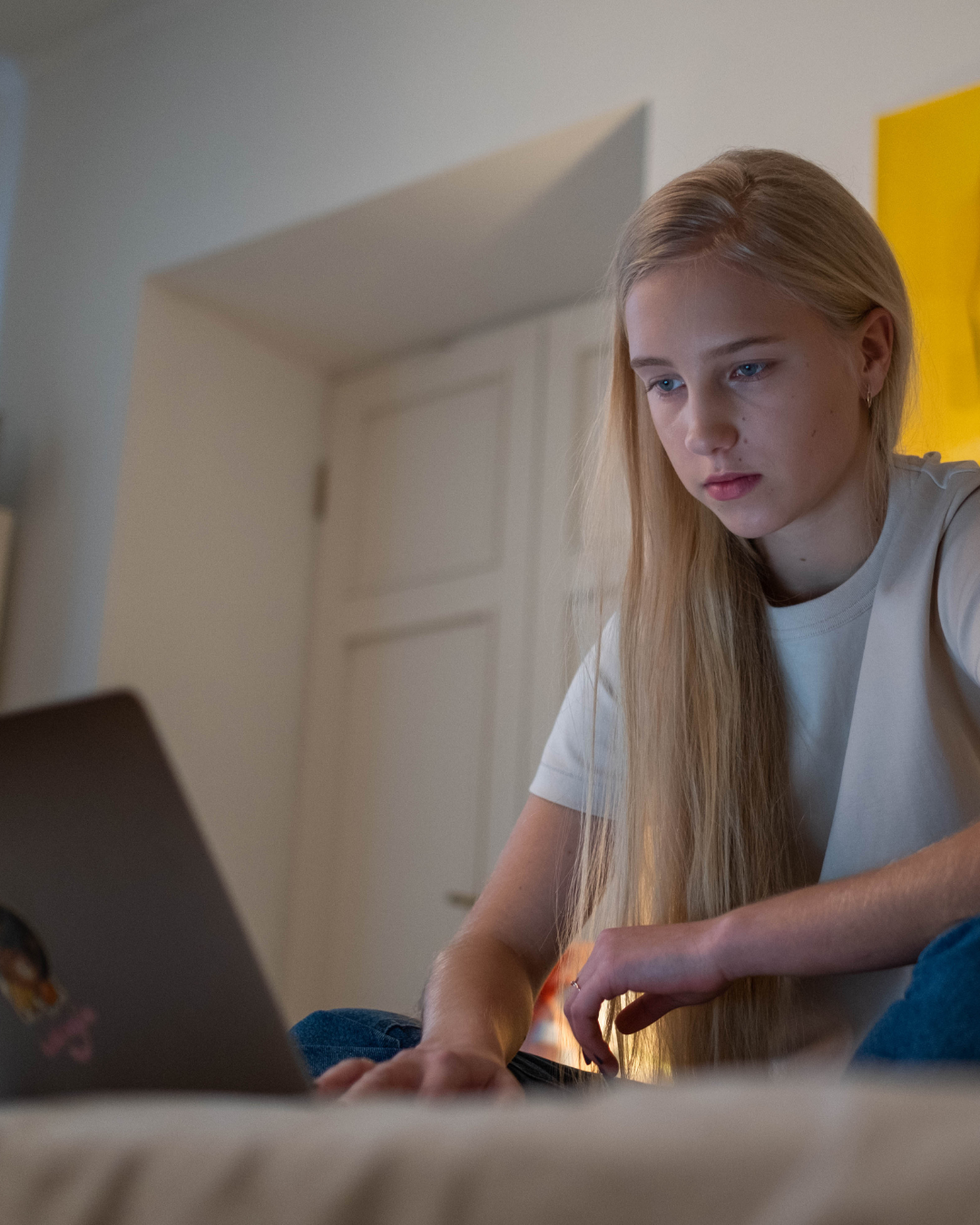 A young woman with long blond hair looking intently at her laptop while sitting on a bed in a cozy room with beige and yellow walls.