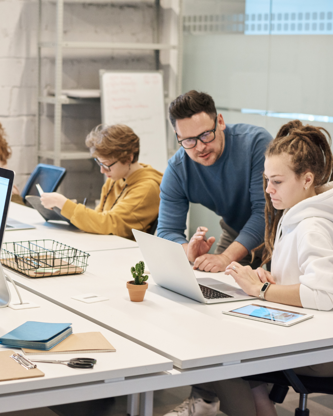 A group of young people working at a long white table in a modern office. One person is standing and assisting another person who is using a laptop, while a third person is focused on a tablet. Items on the table include a small potted cactus, notebooks, scissors, and a wire basket.