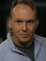 Close-up of a middle-aged man with short hair, wearing a light blue button-up shirt, looking directly at the camera.