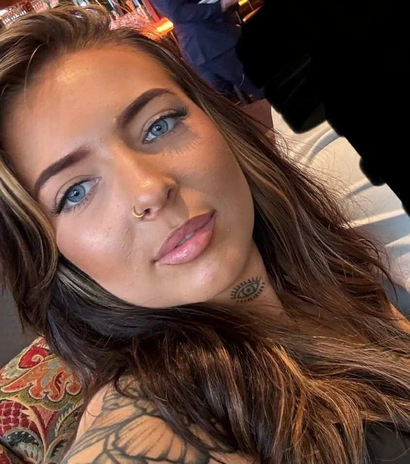 A woman with long dark hair, makeup, and earrings, smiling at the camera, showing a tattoo on her upper arm.