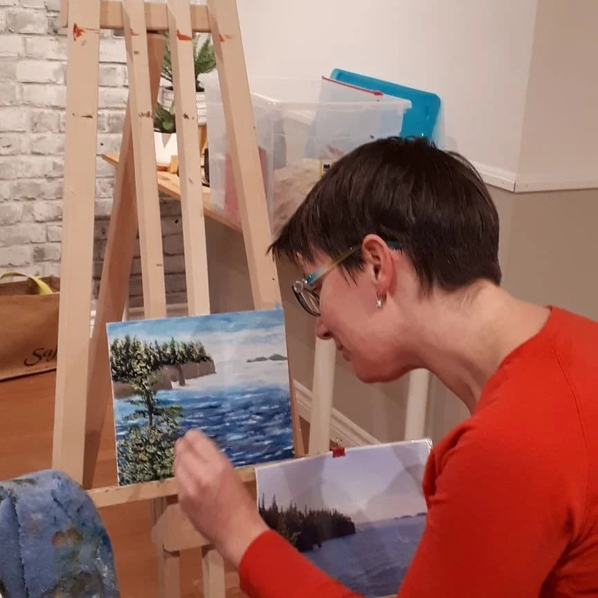 An artist painting landscape scenes of the outdoors, featuring water, trees, and sky, on canvases placed on small easels in an indoor studio.