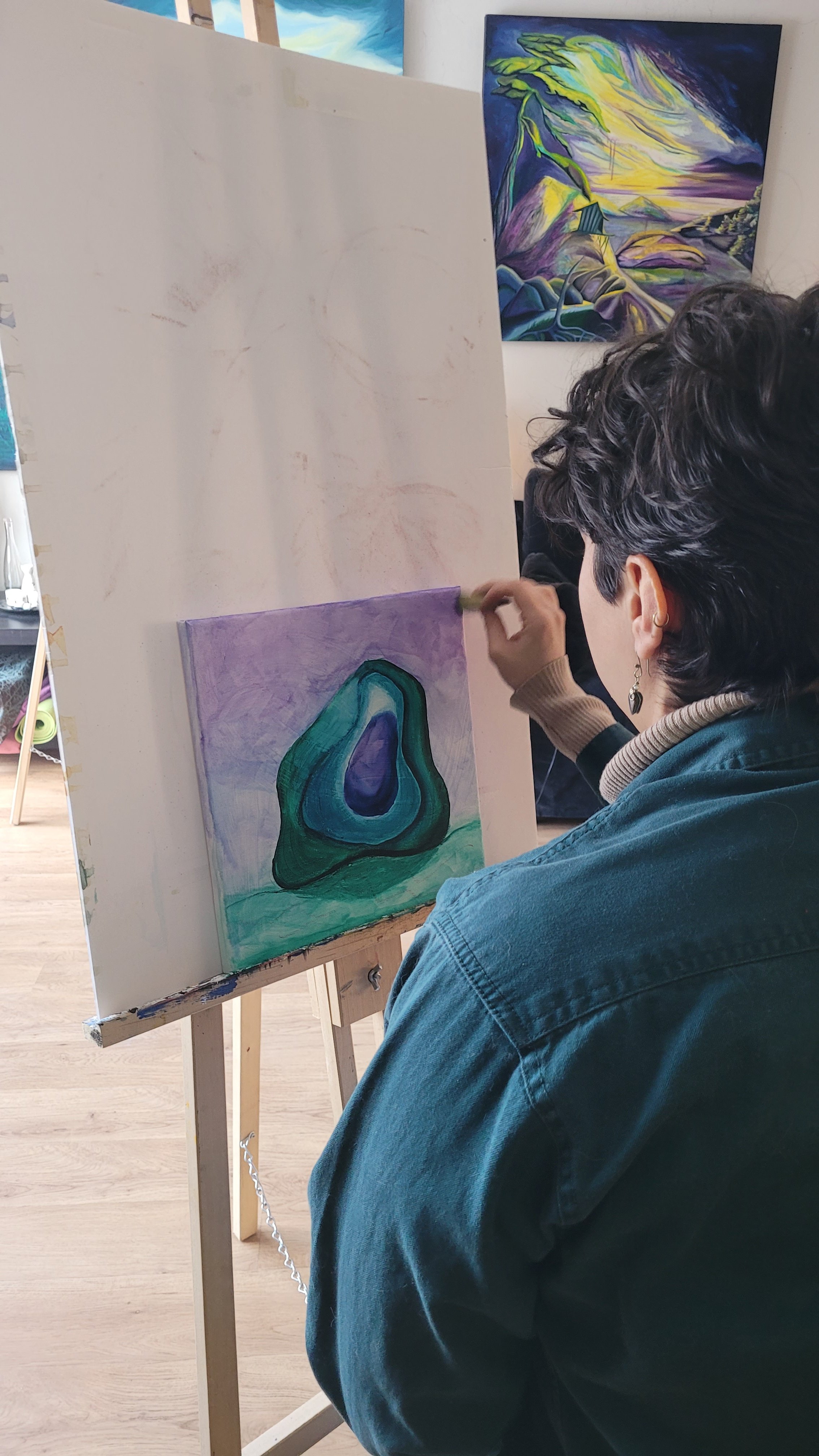 A woman painting with a brush on a canvas features a colorful abstract shape with green, purple, and blue shades, in an art studio with paintings on the wall.