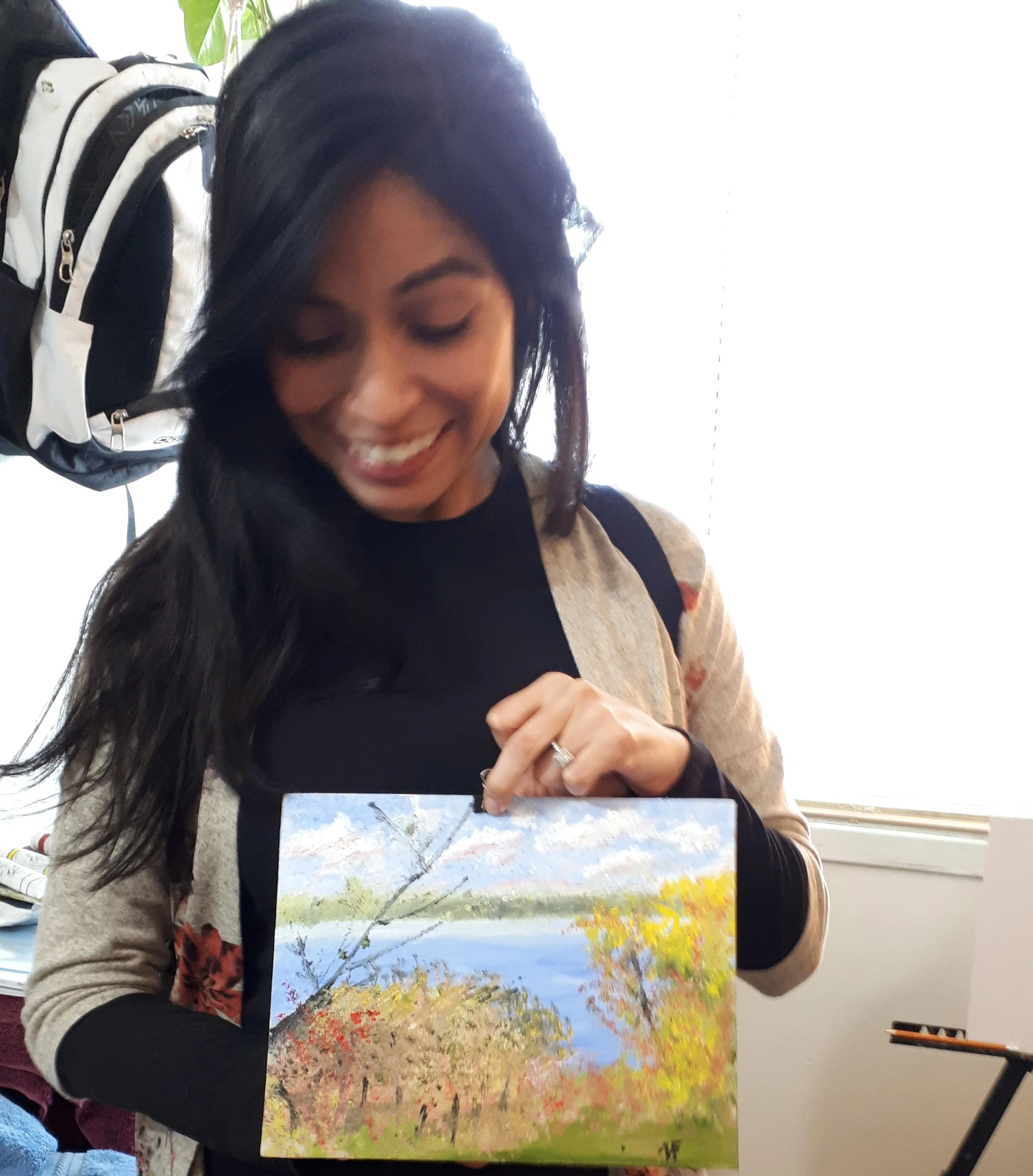 A woman with long dark hair smiling and looking down, holding a colorful landscape painting of trees and a river.