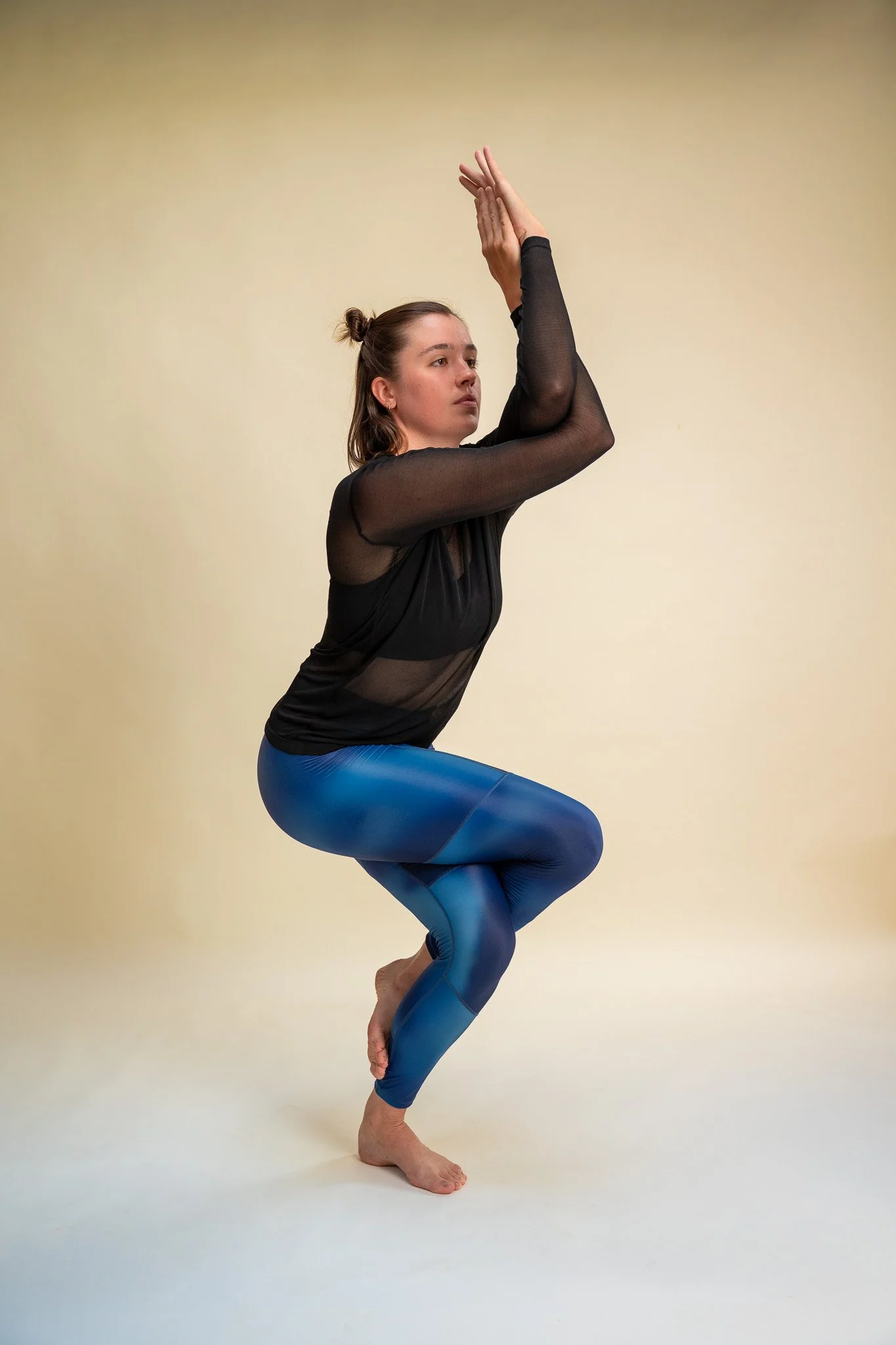 En kvinde, der står i en yoga-positiv med hænder hævet over hovedet og håndflader mod hinanden, iført en sort gennemsigtig langærmet top og blå yoga tights på et neutral baggrund.