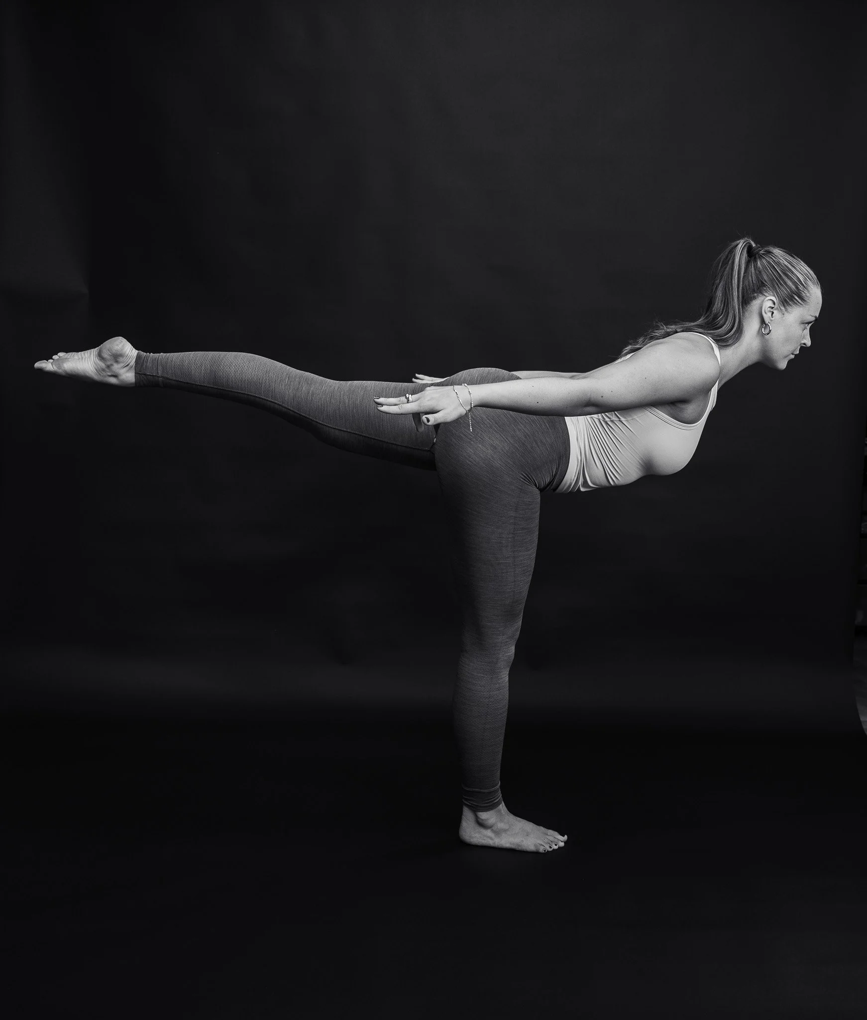 Sort-hvid foto af en kvinde i yoga-stilling, balancerer på ét ben med den anden arm udstrakt bagud og den modsatte arm fremad, mod en sort baggrund.